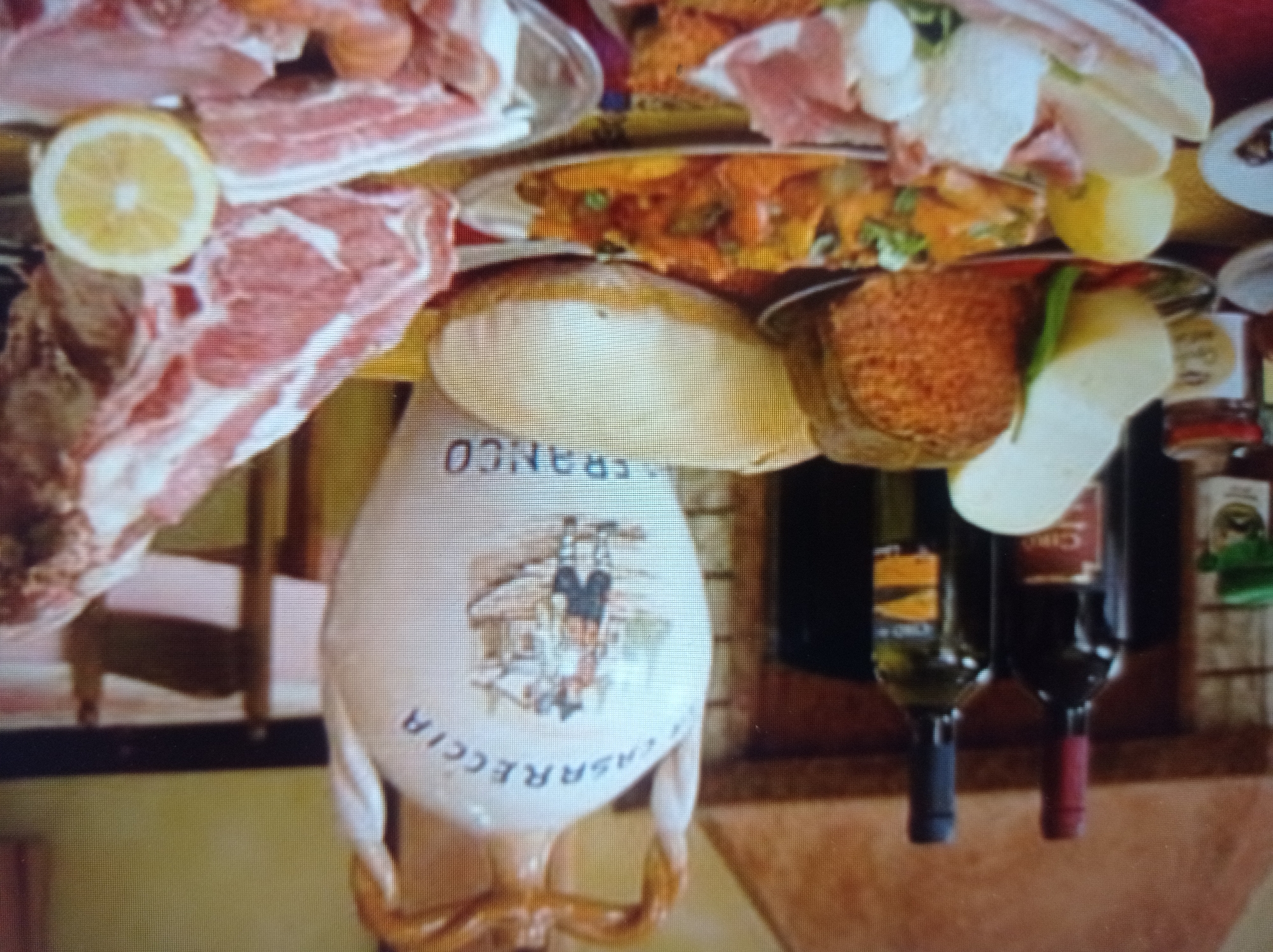 The image depicts a table laden with food and drinks, possibly in a restaurant setting. Dominating the scene is a large, white ceramic jug with a rustic painting and text, likely a decorative wine jug. To the left, two bottles of wine stand tall, one with a red cap and the other with a blue cap. Beside them, various cheeses and cured meats are arranged on plates, suggesting a charcuterie or antipasto selection. A generous portion of raw meat, possibly beef or lamb ribs, is visible on the right side of the frame, garnished with a slice of lemon. In the background, a fireplace or hearth can be discerned. The overall impression is one of abundance and a celebratory meal. The presence of the restaurant chair suggests an indoor dining area. There are no people visible in the image. The provided information about the city of Mukachevo, Ukraine, is not visually verifiable from the image itself and cannot be included in a description based solely on the image content.
