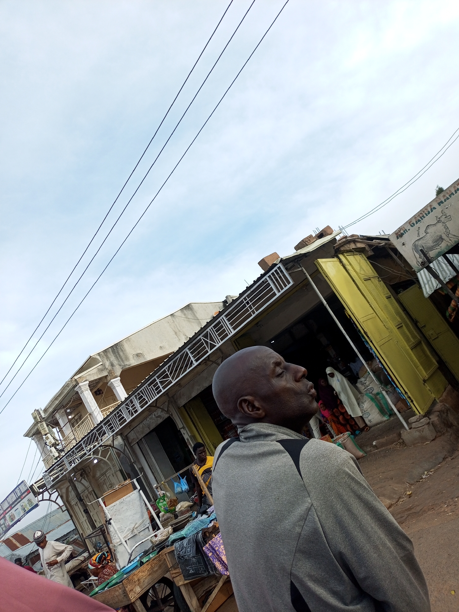 The image captures a street scene in Tempure, Nigeria, with a focus on a bald man in the foreground wearing a grey jacket with black accents. He is looking upwards and to the right, with his mouth slightly open. Behind him, a bustling market environment unfolds.
Several market stalls are visible, one with bright yellow shutters and another with a sign featuring a drawing of a cow and the text "M. GARBA RAMA". In the background, other individuals can be seen, including a person in a white head covering and a man wearing a yellow and black striped shirt. Various goods are displayed, including bags of what appear to be grains or other commodities, and fabric items. A cart with wooden planks and wheels is present, laden with goods. Power lines crisscross the sky above the market. The overall environment suggests an active commercial area in an urban setting.
