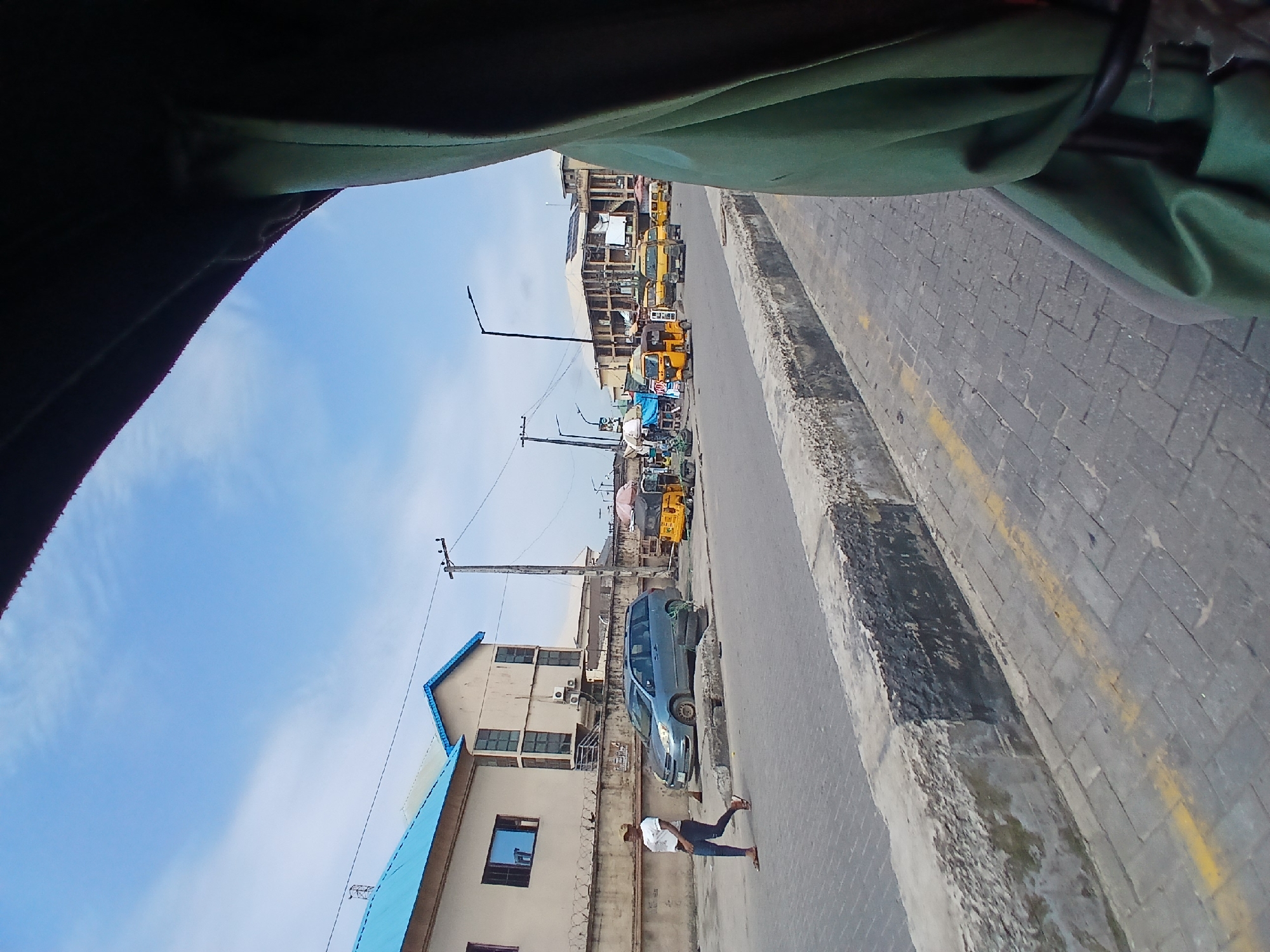 In Aganu, Nigeria, a woman in a white t-shirt and jeans walks across a paved street. She is wearing a tan cap. She walks from left to right across the frame. Beside her, a blue minivan is parked facing left. It is a bright, sunny day with a blue sky and scattered clouds. Several yellow auto rickshaws, or "keke napeps," are visible on the street, along with other vehicles and market stalls. The buildings in the background appear to be a mix of residential and commercial structures. The foreground shows the edge of the street with a concrete curb and yellow painted lines. The image is taken from a perspective that suggests the photographer is inside a vehicle.
