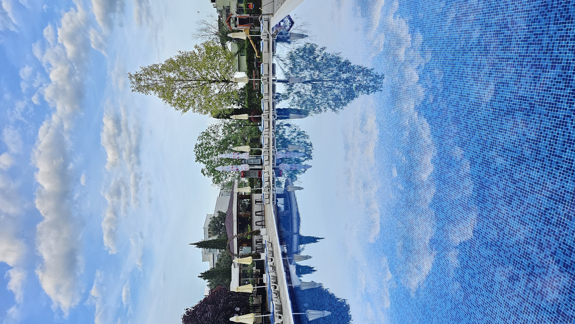 The image shows a swimming pool in Varna, Bulgaria. The pool is filled with water and has a blue tile surface. The sky is reflected in the water, along with the surrounding trees, buildings, and umbrellas. The image is taken from a low angle, giving the viewer a sense of being in the water. There are no people visible in the image.