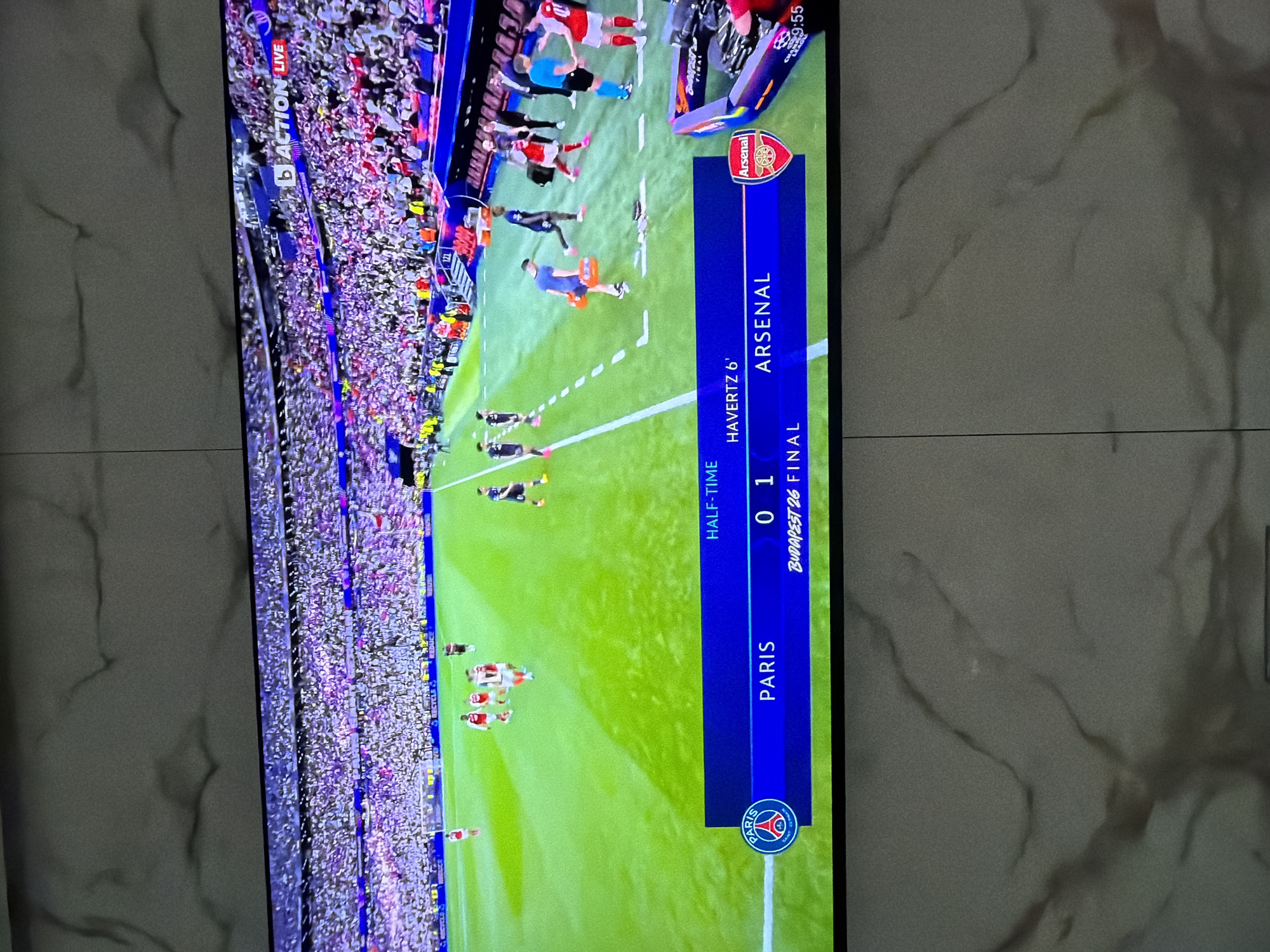 The image shows a television screen displaying a soccer match between Paris Saint-Germain (PSG) and Arsenal. The score is 0-1 at halftime, with Havertz scoring for Arsenal in the 6th minute. The match is the Budapest 26 final and is being broadcast live on "Action Live." The TV is positioned against a wall covered in grey textured tiles. The time displayed in the upper right corner of the screen is 9:55.