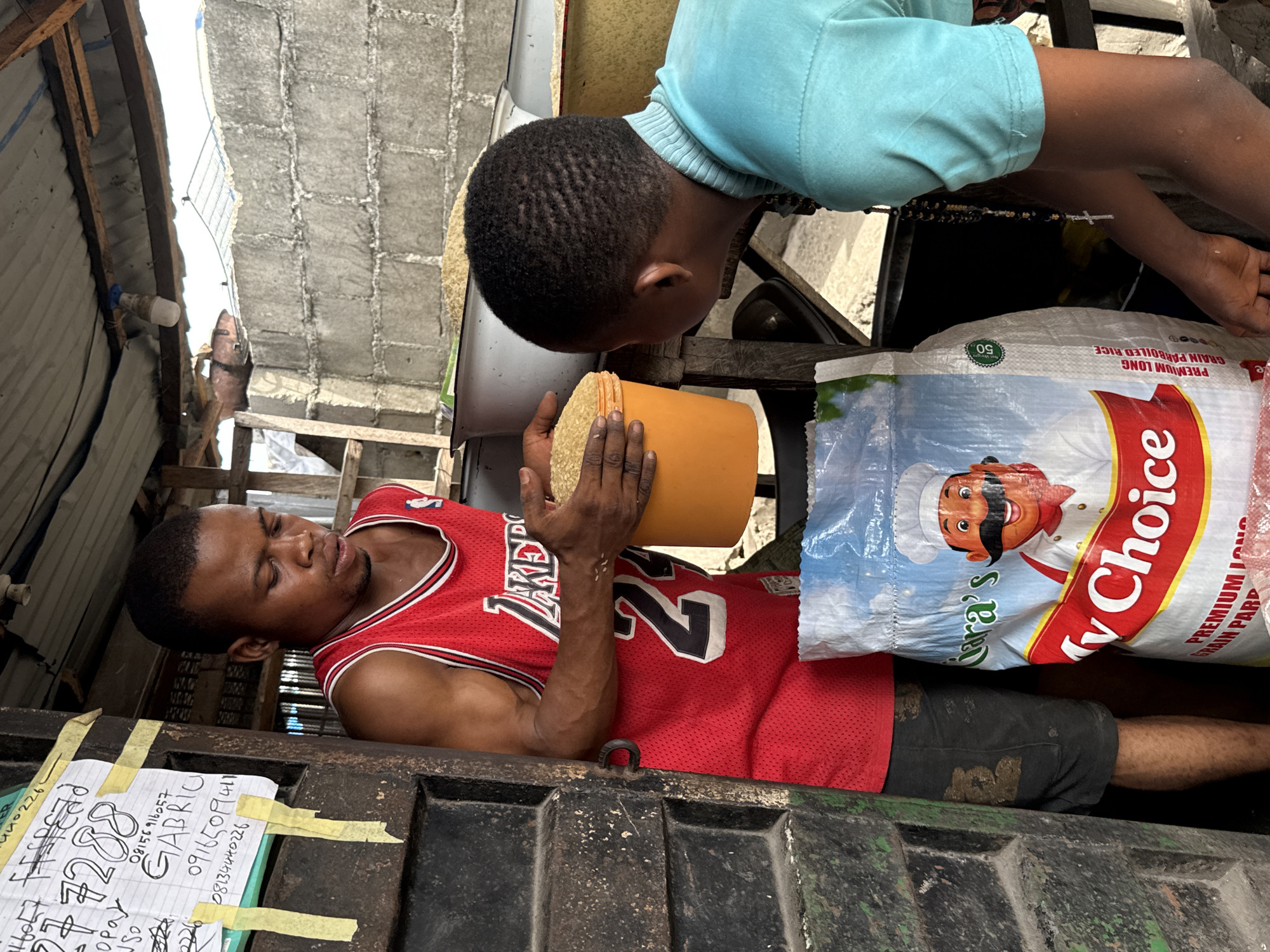 This image, taken in Ado, Nigeria, captures two males engaged in handling grains, likely rice, within a semi-enclosed, utilitarian space. The scene appears to be inside a market stall, a storage area, or the back of a vehicle, characterized by rough, dark metal surfaces and simple construction elements like concrete block walls, wooden beams, and corrugated metal roofing.
An adult man, wearing a red basketball jersey with "LAKERS" and the number "22", is positioned on his back, holding an orange plastic container filled with light-colored grains. His arms show visible musculature, indicative of physical activity. A younger male, dressed in a light blue long-sleeved shirt, is leaning over, observing the grains in the container. A necklace with a small silver cross pendant is visible on his arm.
Next to the adult man lies a large white sack of "My Choice PREMIUM LONG GRAIN PARBOILED RICE," marked with "50," likely indicating its weight in kilograms. The sack features a smiling chef character.
Taped to a dark metal surface in the foreground is a piece of paper displaying handwritten text. Visible details include numbers "140226," "277288," and "180," along with "OP Opay," the name "GABRIO," and several phone numbers: "08156916057," "09165099411," and "08134440226." These details likely pertain to sales, contacts, or financial transactions.
The setting includes an exposed LED light bulb under the corrugated roof. Both individuals appear focused on the task at hand, with the adult man's eyes slightly closed or squinted in concentration. The lighting suggests it is daytime.