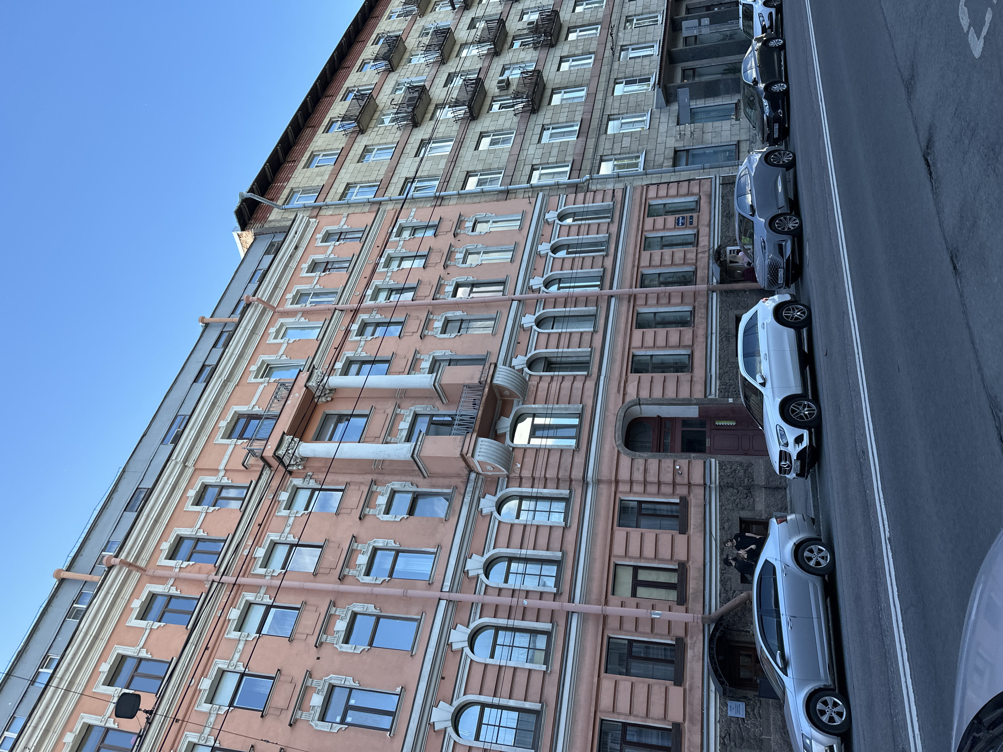 This image captures a street scene in Saint Petersburg, Russia, featuring two distinct apartment buildings side-by-side. The building on the left is a salmon-pink, multi-story structure with ornate architectural details around its windows and balconies. The building on the right is a lighter-colored, possibly stone or brick, structure with a repeating pattern of balconies with black railings. The street in front of the buildings is paved and lined with parked cars. The sky above is clear and blue. Several cars are visible parked along the street. On the sidewalk in front of the pink building, two individuals, dressed in dark clothing, are standing and interacting. The overall atmosphere is one of an urban residential setting under a clear sky.