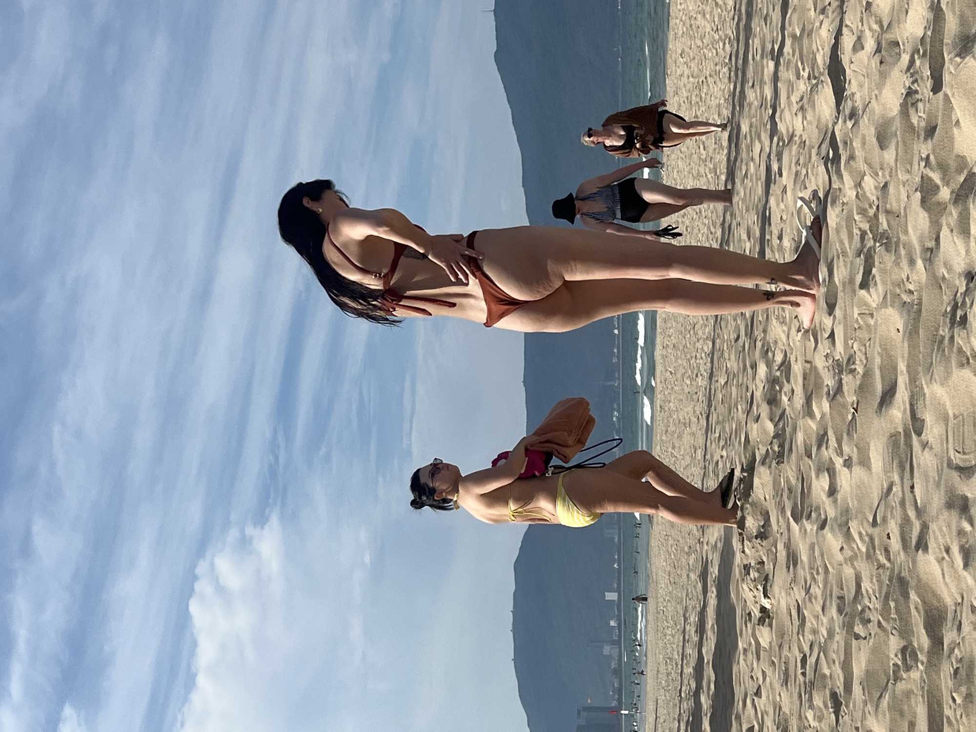 A wide-angle, eye-level, outdoor photograph captures a sunny day at My Khe Beach in Da Nang, Vietnam. The foreground is dominated by textured sand, with footprints and light shadows from the sun. In the mid-ground, several individuals are present. The most prominent subject is a tall woman in a dark red bikini, facing away from the camera and looking down, her hand on her hip. To her left and slightly further back, a woman in a yellow and white striped bikini walks away from the camera, holding a towel and a pink bag. Further in the background, two other women are visible: one wearing a black and white striped swimsuit and a dark hat, and another in a brown cover-up over a dark bikini. They are walking towards the right side of the frame. Scattered across the beach, people can be seen in the distance, wading in the ocean and walking along the shoreline. The ocean stretches to the horizon, with small waves breaking. In the background, a mountain range rises behind a city skyline with buildings. The sky is a clear blue with scattered white clouds.