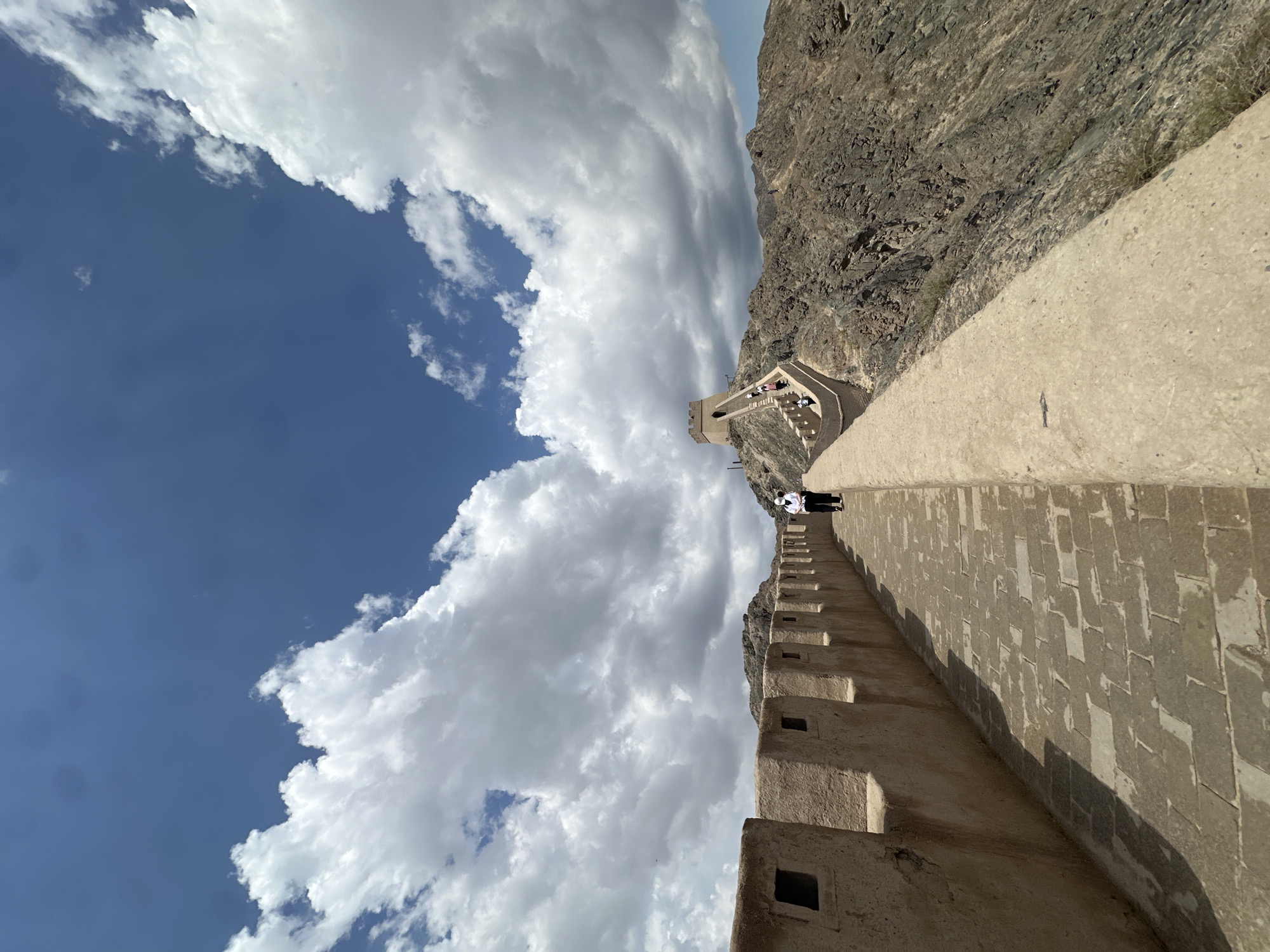 A wide-angle, eye-level, outdoor photograph captures a section of the Great Wall of China, likely near Jiayuguan, China, under a bright, partly cloudy blue sky. The wall, constructed from tan-colored bricks and stone, snakes along a rugged, rocky mountainside. The walkway on the wall is paved with rectangular stones. Along the top of the wall are crenellations, with square openings visible.
In the foreground, a person wearing a white shirt, dark pants, and a white face mask walks away from the camera along the wall. Further in the distance, the wall ascends a steep incline towards a fortified watchtower situated atop a rocky peak. Several other individuals can be seen on the wall pathway and climbing towards the watchtower. The surrounding landscape is characterized by barren, rocky hills and sparse vegetation. The lighting suggests it is daytime, with the sun casting shadows on the pathway.