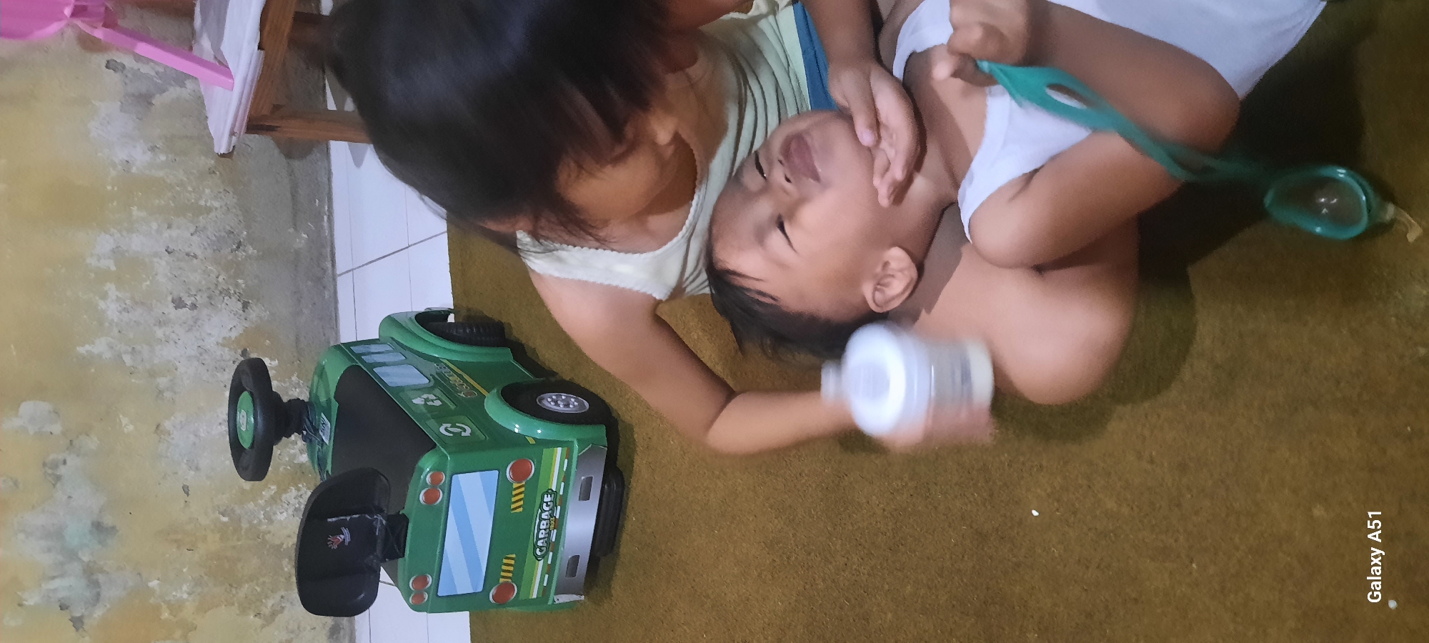 In Jatitengah, Indonesia, two young children are on a carpeted floor. The older child, a girl with dark hair, is leaning over the younger child, a boy, who is lying on his back. The girl is wearing a light-colored sleeveless shirt, and the boy is wearing a white sleeveless undershirt. The girl is holding a white baby bottle with her right hand and has her left hand near the boy's chin. The boy is looking upwards with his mouth open and eyes partially closed. A teal-colored spoon-like object is held by the boy. To the left of the children is a green toy garbage truck. The background shows a textured, yellowish wall and a section of white tiled floor.