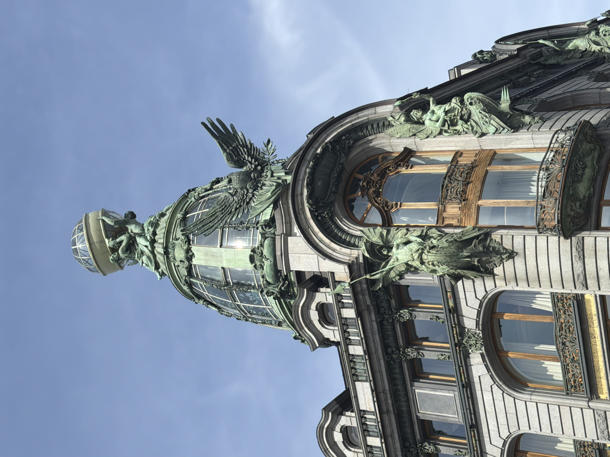 The image depicts a corner view of the Singer House (also known as the Dom Knigi or House of Books) in Saint Petersburg, Russia. The building is oriented vertically in the image. The architectural style is Art Nouveau, with elaborate ornamentation. Key features include a glass dome topped with a sculptural group and a prominent eagle sculpture with outstretched wings on the side of the dome. The building facade exhibits light-colored stone or stucco, with multiple arched windows accented by decorative ironwork and bronze sculptures. The sky in the background is light blue with minimal clouds.