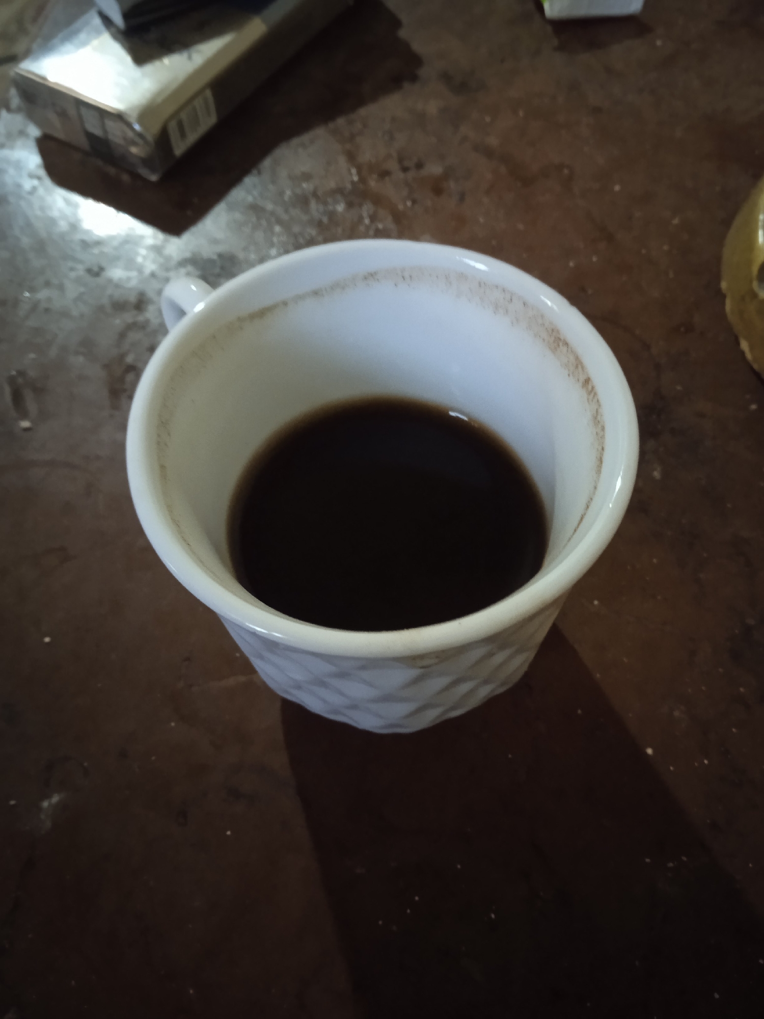 A close-up, high-angle shot shows a white mug filled with dark liquid, presumably coffee, placed on a dark, textured surface. The mug has a quilted pattern on its exterior and a faint ring of residue is visible along the inside rim. In the upper left corner of the frame, a portion of a cigarette pack is visible. The background is out of focus, but appears to be a table with some scattered light-colored specks. The scene is lit, creating shadows. There are no people present. The image does not provide any specific information about the location of Aznvadzor, Armenia.