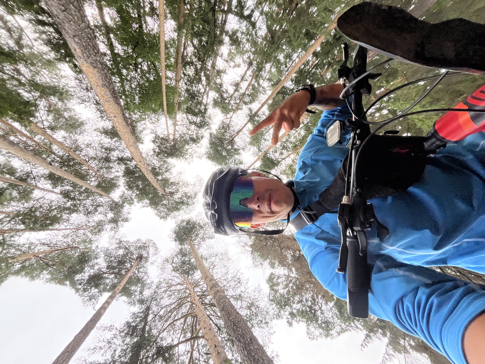 A first-person perspective shot captures a person wearing a black helmet, iridescent cycling glasses, and a bright blue long-sleeved cycling jacket. The individual is holding the handlebars of a bicycle, and their left hand is raised in a peace sign. The scene is set within a forest with tall pine trees reaching towards an overcast sky. The image appears to be taken on a trail within the city of Chaikovsky, Russia, given the distinctive coniferous forest characteristic of the region.