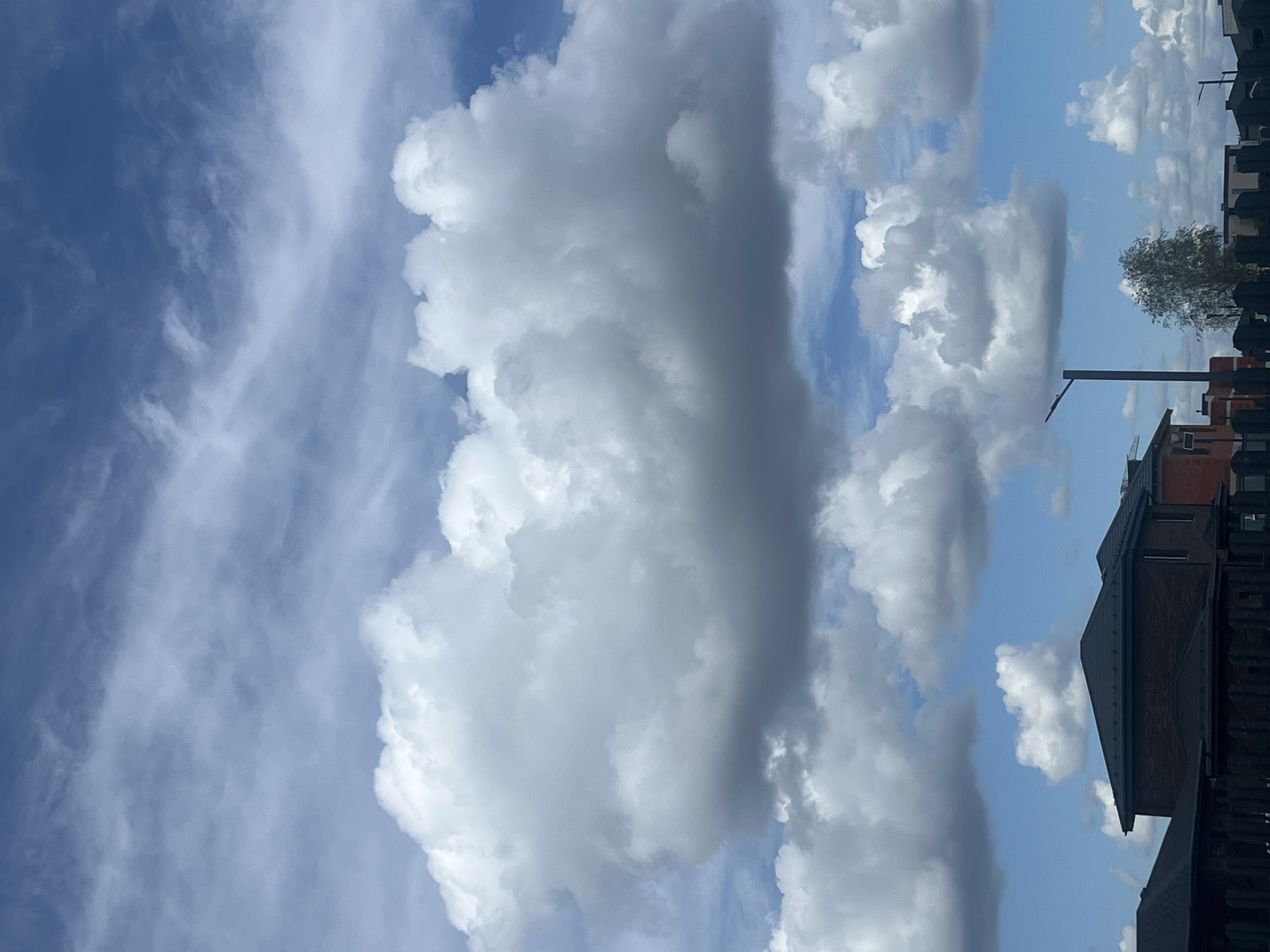 The image shows a sky filled with fluffy white and grey clouds, with patches of blue sky visible. In the lower part of the frame, several modern brick houses with dark roofs are visible. A street lamp stands to the right of a tree. A dark fence runs across the bottom of the image. The scene appears to be in a suburban area.