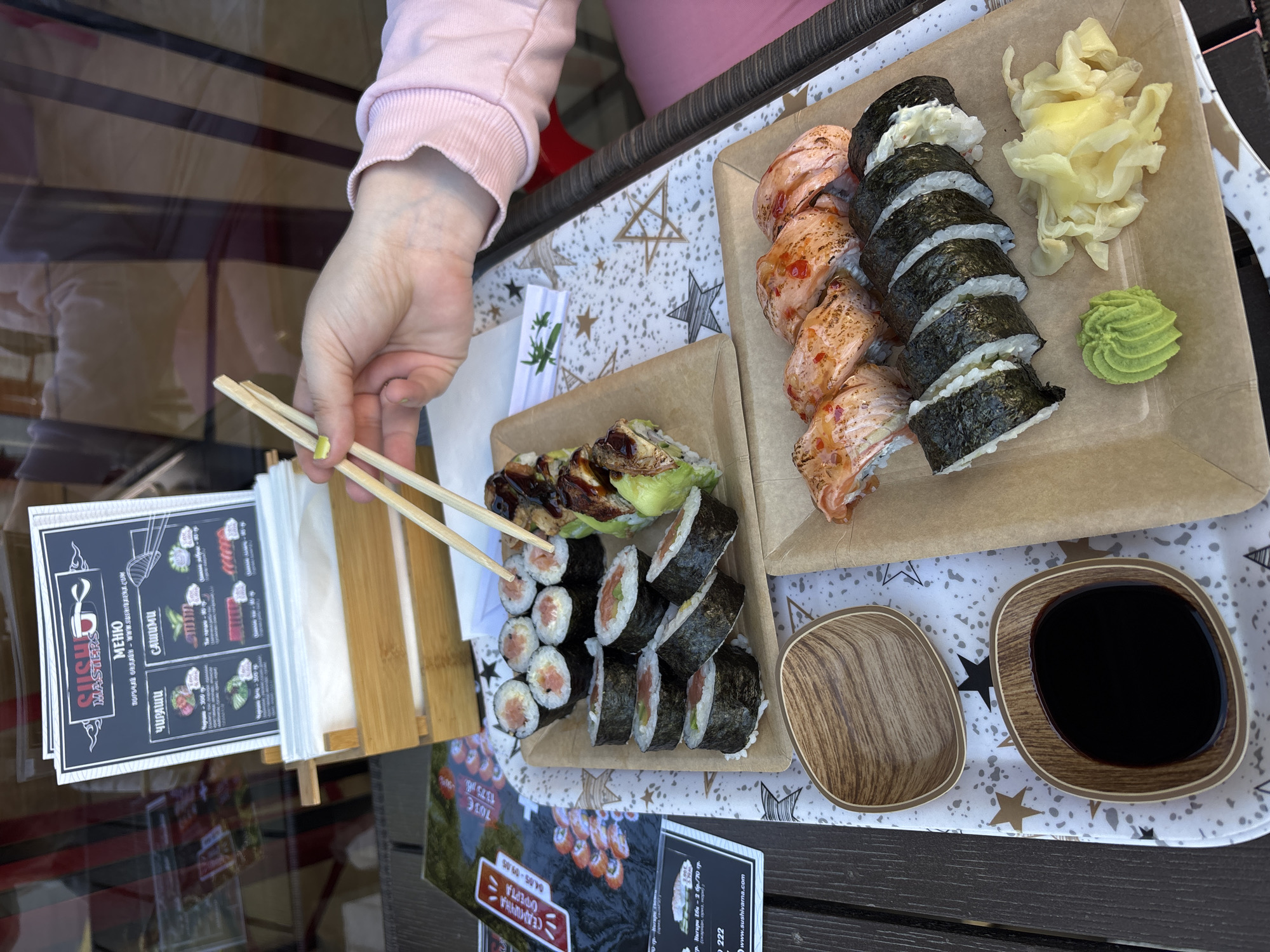 The image shows a tray of sushi at a restaurant or outdoor dining setting. A hand with chopsticks is reaching for sushi. Two cardboard trays hold different kinds of sushi rolls, pickled ginger, and wasabi. Two small wooden bowls, one empty and the other filled with soy sauce, are present. A menu from Sushi Masters is visible. The tray with the sushi sits on a table with a star-patterned design. Information on the menu and a nearby sign suggest the location is in Varna, Bulgaria.