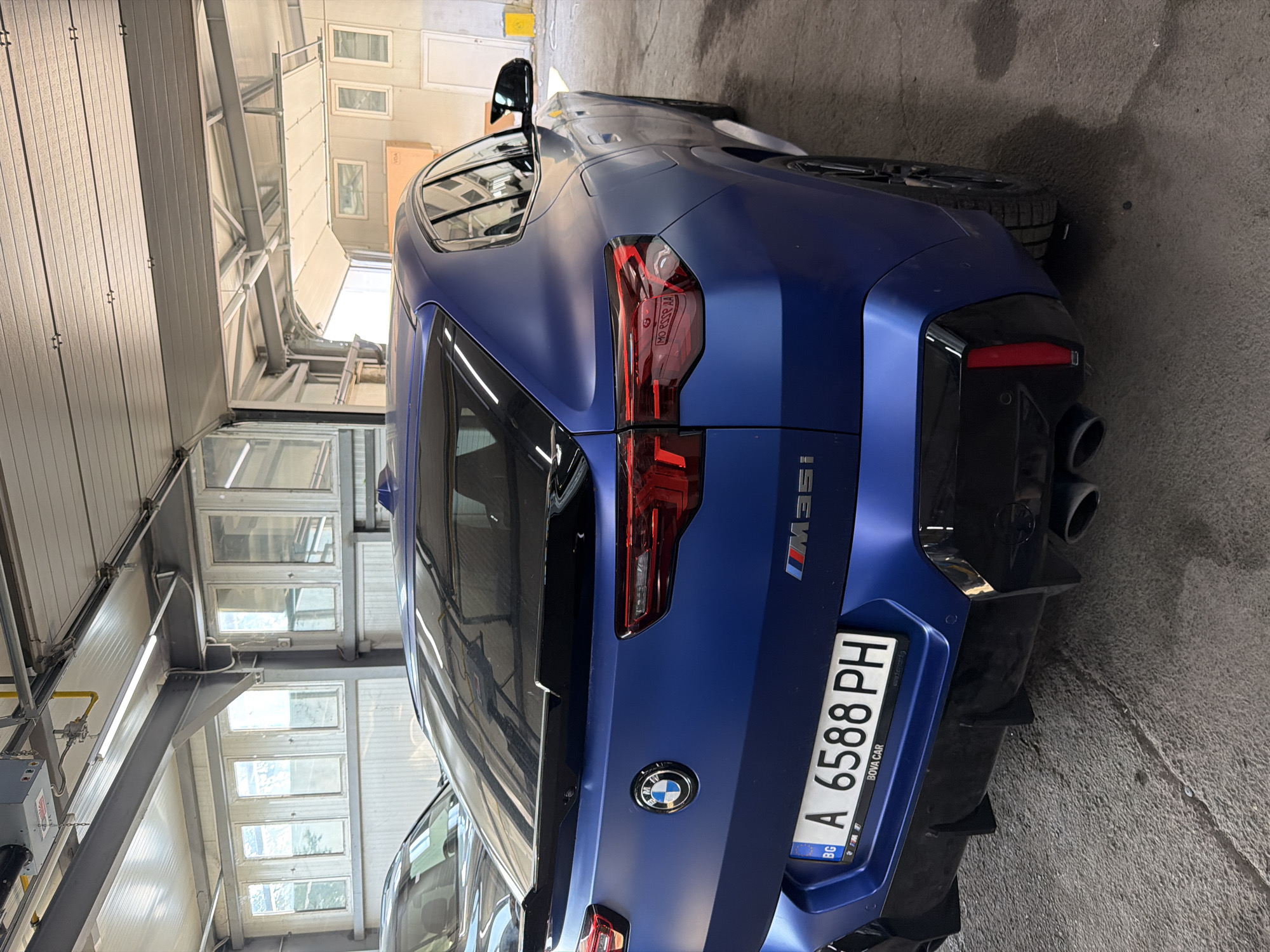 The image displays a matte blue BMW X2 M35i, captured from its rear-right side, parked indoors within what appears to be a garage or automotive workshop. The car features a sporty aesthetic with an "M35i" badge on the tailgate, distinctive LED taillights, and a rear bumper incorporating a black diffuser with prominent quad exhaust tips. A Bulgarian license plate, "A 6588 PH", is clearly visible, signifying registration in Burgas, Bulgaria, and the frame includes the text "BOVA CAR". The BMW logo is also present on the tailgate.
The setting consists of light-colored, insulated panels resembling garage doors or walls on one side, with visible metal structural beams and multiple rectangular windows providing natural light. The floor is concrete. The overall scene suggests a clean, professional automotive environment, possibly a service center, dealership, or private garage. The car is stationary, with no specific activity taking place. The lighting indicates it is daytime, but no external weather conditions are visible.