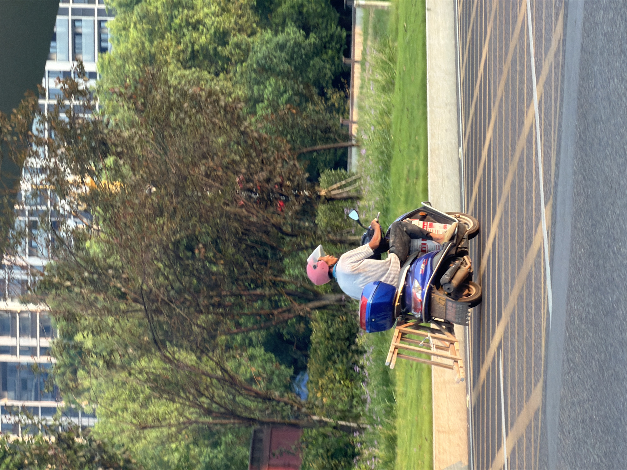 The image shows a person riding a blue scooter on a street in Jiangjin, China. The person is wearing a pink helmet and a striped shirt. The scooter has a blue storage box on the back and a wooden ladder attached to it. The scooter is moving from left to right. In the background, there are trees and buildings. The street has lane markings. The lighting suggests it is late afternoon or early evening.