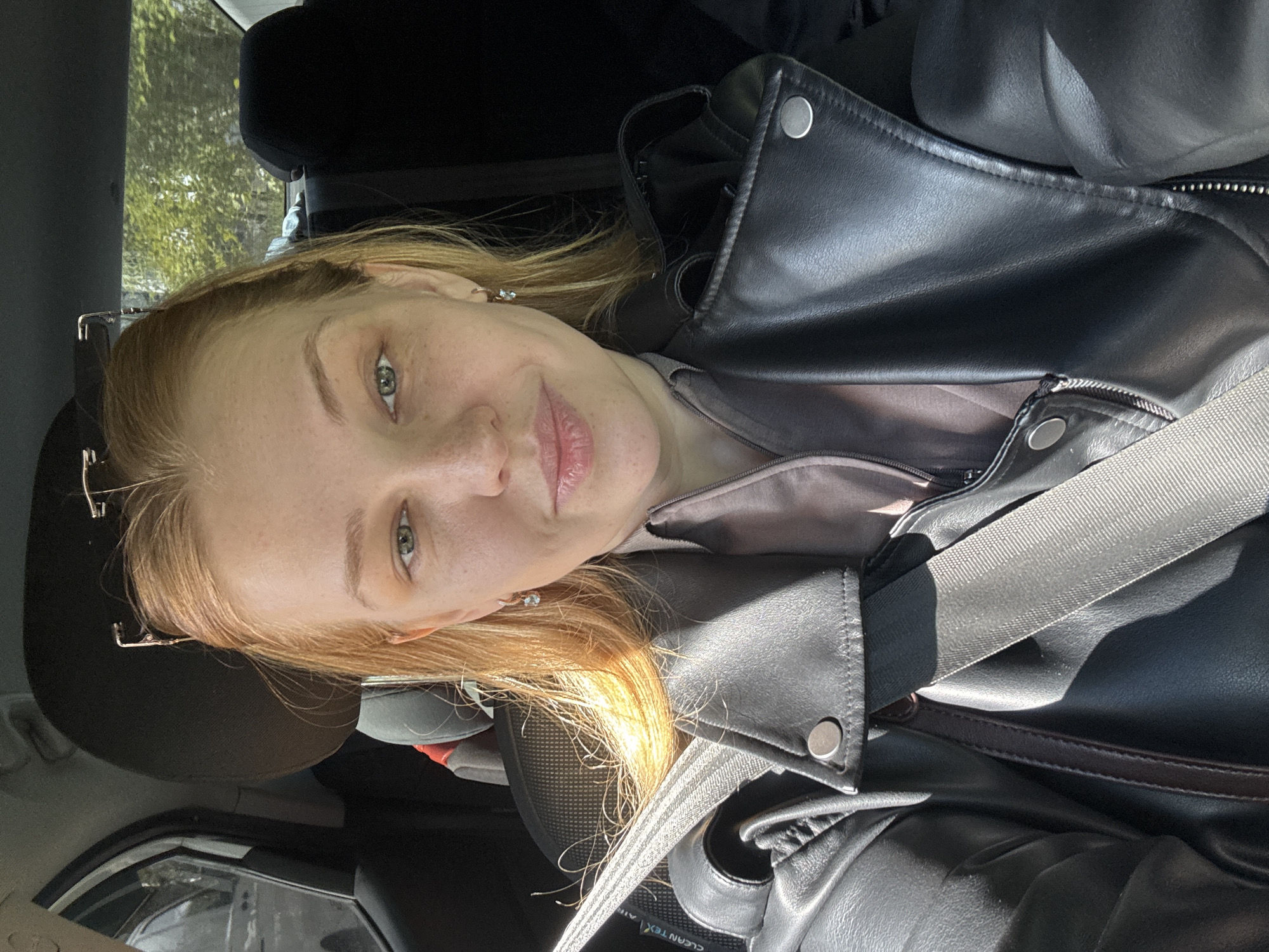 A selfie of a woman with light skin and reddish blonde hair, presumably inside a car. She is wearing a black leather jacket over a light brown top and has a seatbelt fastened. The interior of the car is visible, with a headrest and parts of the dashboard and windows. The location is unknown.