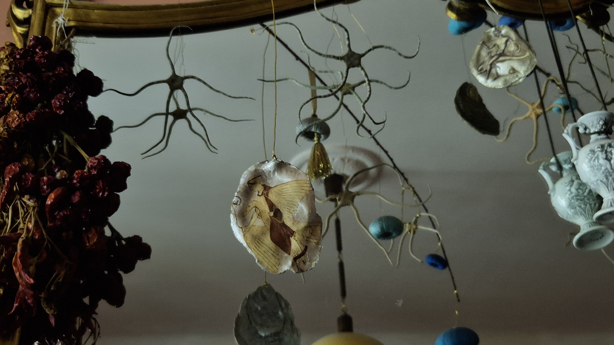 The image depicts a collection of eclectic decorative items hanging from what appears to be a mirror frame, reflecting a light-colored ceiling. Prominently featured on the left is a bunch of dried red chili peppers, casting a dark silhouette. Suspended from fine threads are various ornaments, including star-shaped dried flora, a light-colored translucent disk with a painted depiction of a winged figure, a golden tassel, and small blue spherical objects. To the right, three small, ornate, pale blue or white vases are visible. The overall impression is one of a carefully curated and somewhat whimsical arrangement of objects. The specific location within Taranto, Italy is not discernible from the image itself.