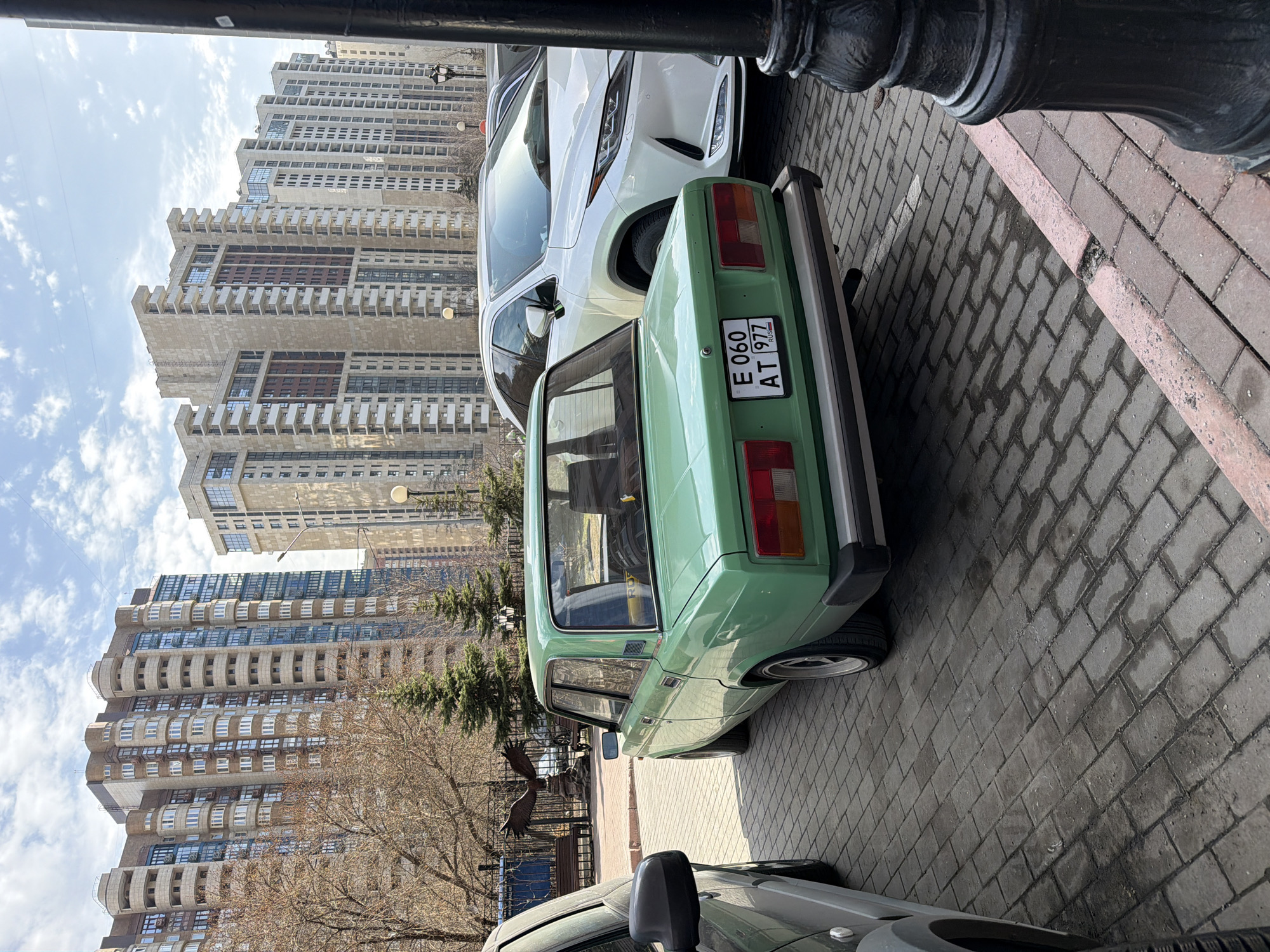 The image depicts a light green Lada parked on a brick-paved street in Moscow, Russia. The license plate reads "E 060 AT 977 RUS." A white SUV is parked in front of the Lada. High-rise residential buildings are visible in the background. The weather is clear with some clouds. A black metal post is in the foreground.