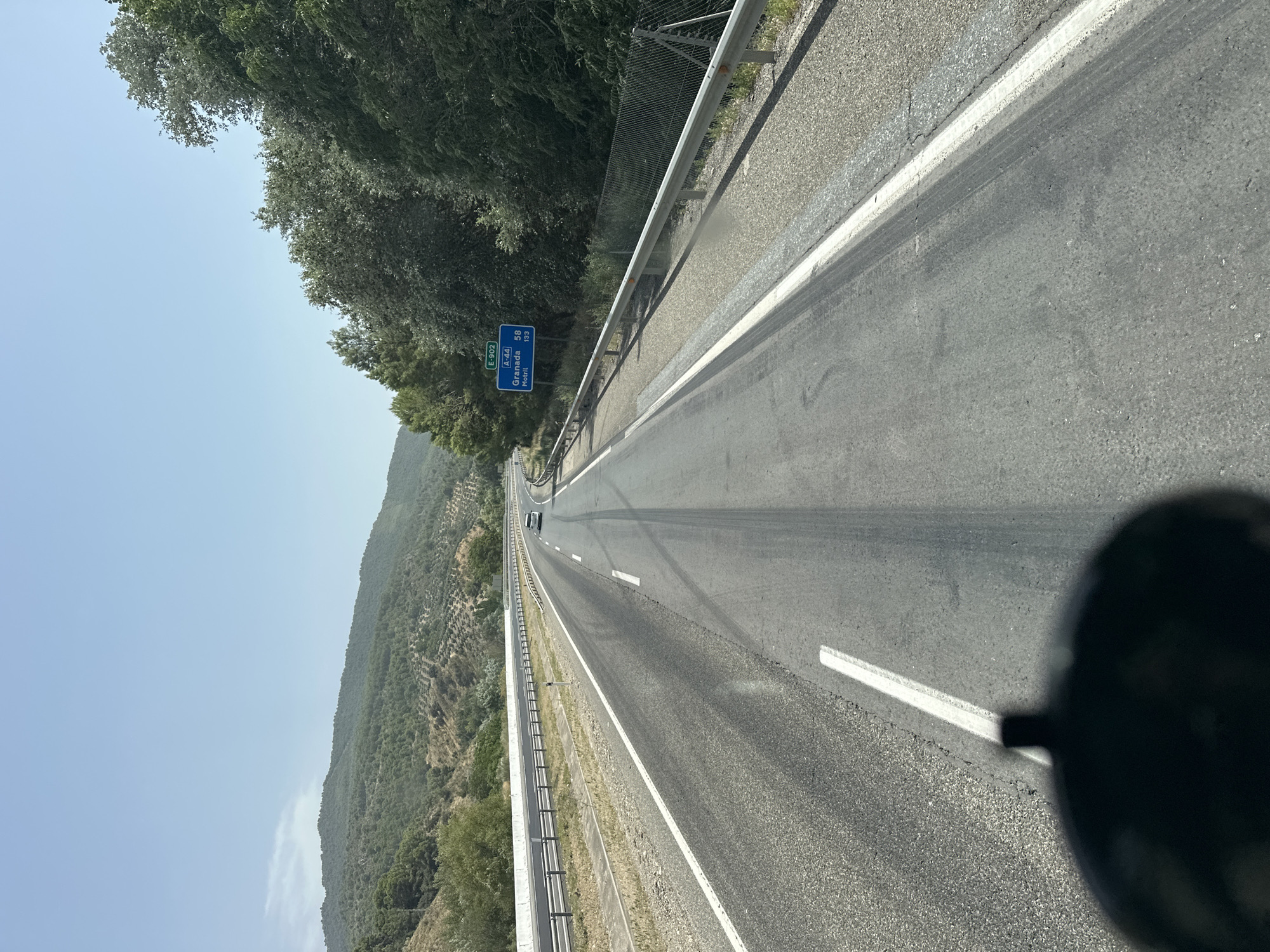 The image captures a forward view from a vehicle on a highway in the vicinity of Cambil, Spain. The road, an asphalt surface, is marked with white lines and tire tracks are visible. To the right, a metal guardrail separates the road from a gravel shoulder and a chain-link fence. Beyond the fence, dense green trees line the road. A blue road sign indicates "E-902" and "A-44" with destinations "Granada 58" and "Motril 133", suggesting the location is on the A-44 highway. In the background, rolling hills covered in vegetation and some scattered olive trees are visible under a clear blue sky. A white car is seen driving ahead on the highway. A dark, shadowed object, likely part of the vehicle from which the photo is taken, is visible in the lower foreground.