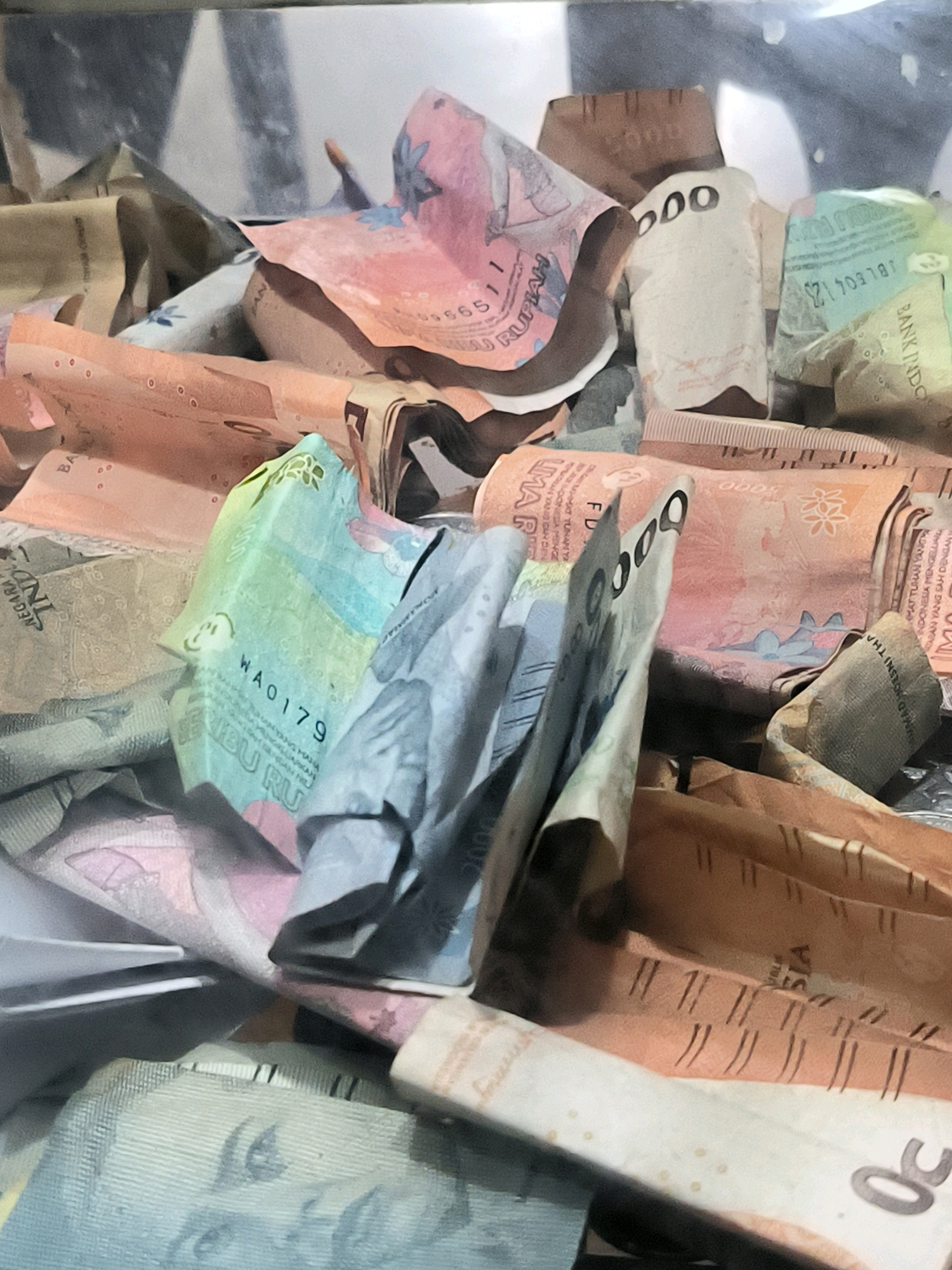 The image shows a close-up of a pile of Indonesian Rupiah banknotes. The banknotes are folded and crumpled, and several denominations are visible. Some of the visible denominations include 5000, 10000, and 50000. The colors of the banknotes vary, with shades of pink, orange, green, and white. The background is out of focus, but appears to be a dimly lit interior space. The image does not provide any specific information about the location in the city of Banda Aceh, Indonesia, as it focuses solely on the currency. There are no people or actions depicted in the image.