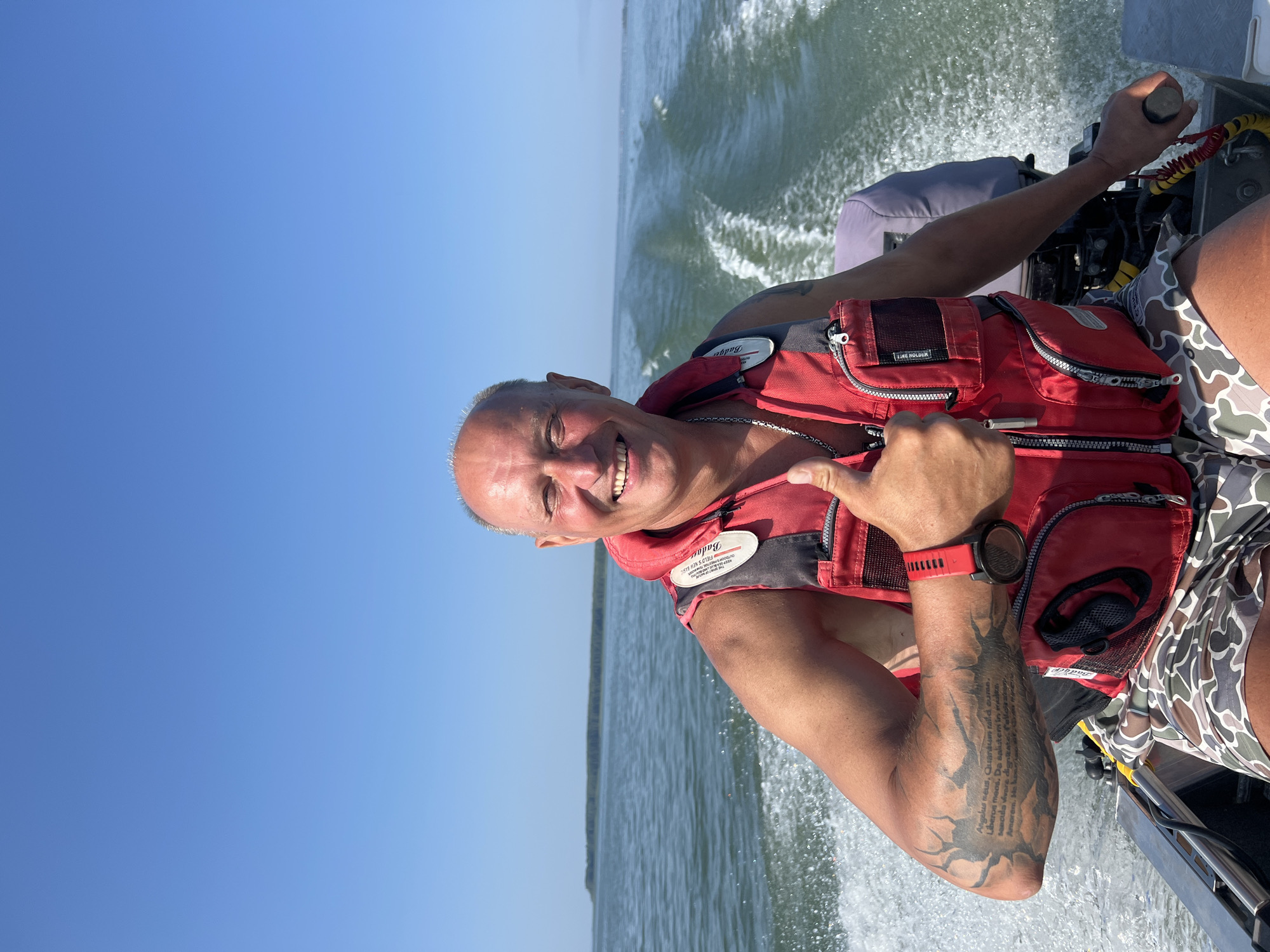 A man wearing a red life vest and camouflage shorts is on a boat. He is smiling and giving a thumbs-up. The boat is moving through the water, creating a wake. The sky is clear and blue, and there is a line of trees in the distance. The location is the city of Beregovoe, Russia.
