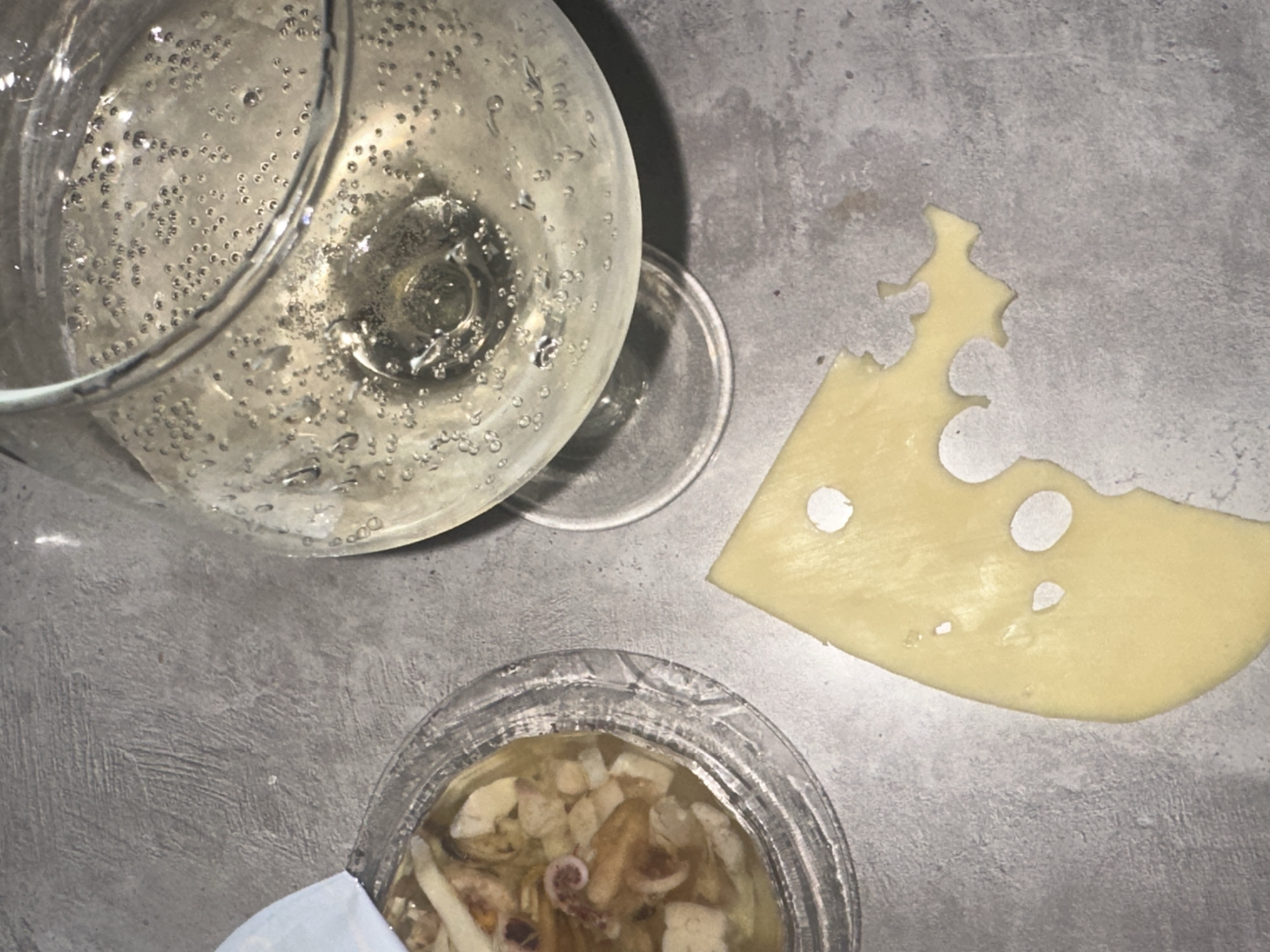 The image presents an overhead view of a gray surface, possibly a countertop or table. A glass of sparkling wine filled with a bubbly, clear liquid occupies the upper left quadrant. Next to it, a piece of Swiss cheese with distinctive holes is situated. In the lower center, a small clear plastic container holds a mixture of seafood, including what appears to be squid and other ingredients suspended in a liquid. The location, Chaikovsky, Russia, is speculative and not verifiable from the visual data alone. There are no people visible, and the only actions implied are the preparation or consumption of food and drink.