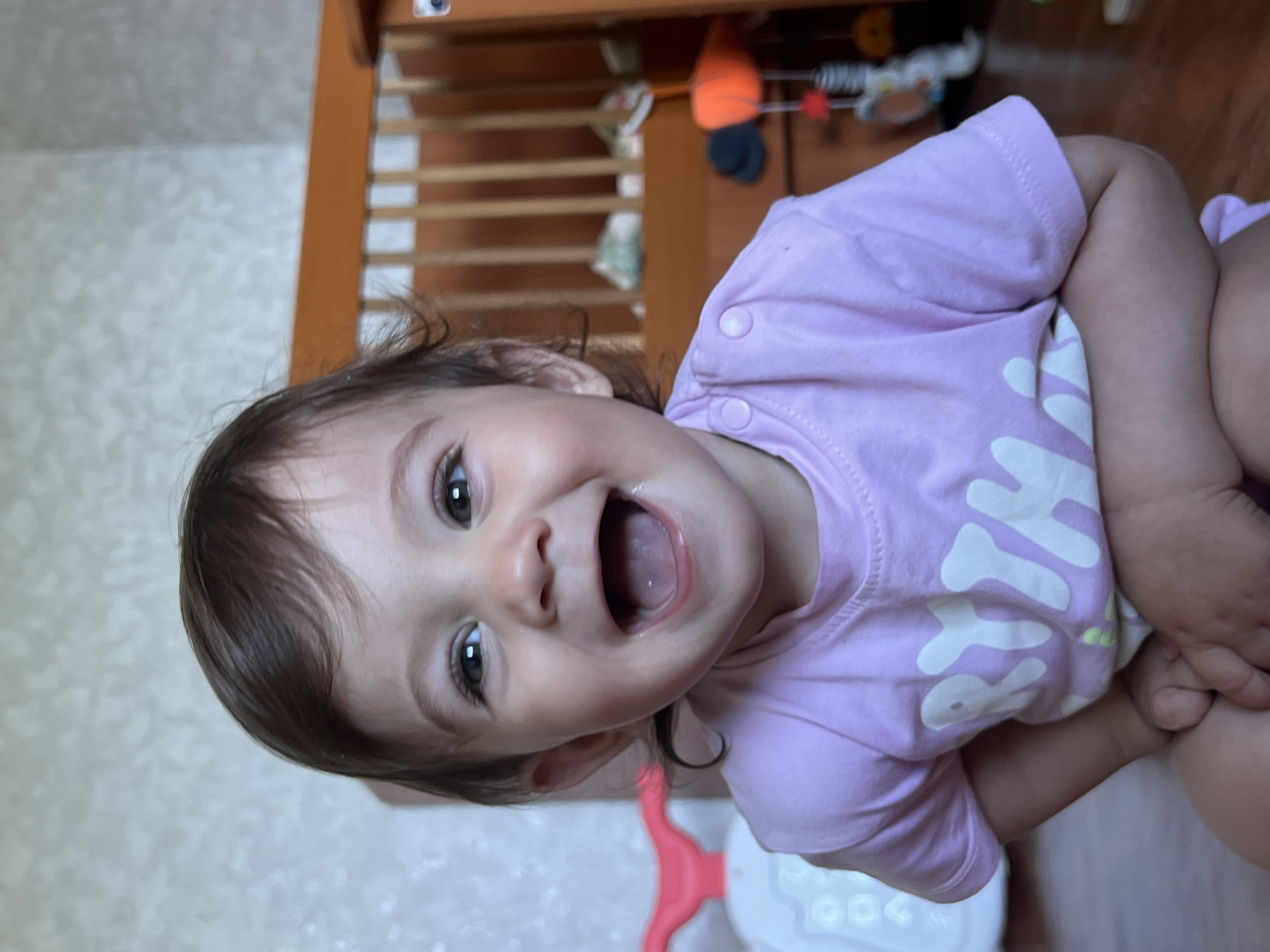 A close-up, eye-level, indoor shot features a laughing baby with dark brown hair and large, dark eyes. The baby is wearing a light purple t-shirt with white lettering. The background consists of a beige, textured wall and a wooden crib. The baby appears to be in a residential setting, possibly a nursery. No specific location in Elena, Bulgaria can be determined from the image.