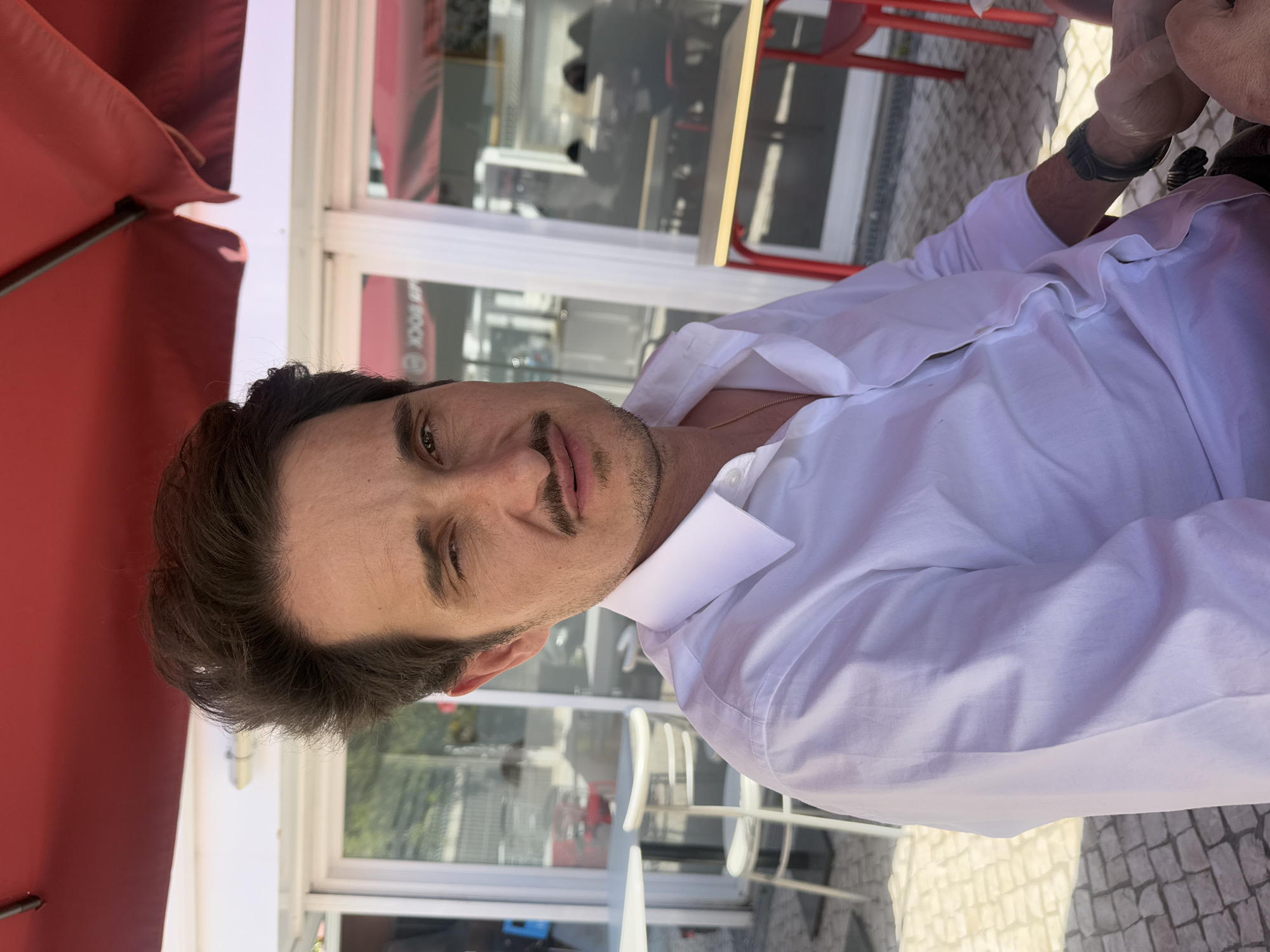 The image shows a man with dark hair and a mustache, wearing a white shirt. He's sitting outdoors at a cafe or restaurant, likely in Cascais, Portugal, as evidenced by the local "OCK" branding visible in the background. A red umbrella is partially visible above him. He appears to be looking at the camera. Other tables and chairs are seen in the background, suggesting an outdoor dining area. The setting seems to be urban.