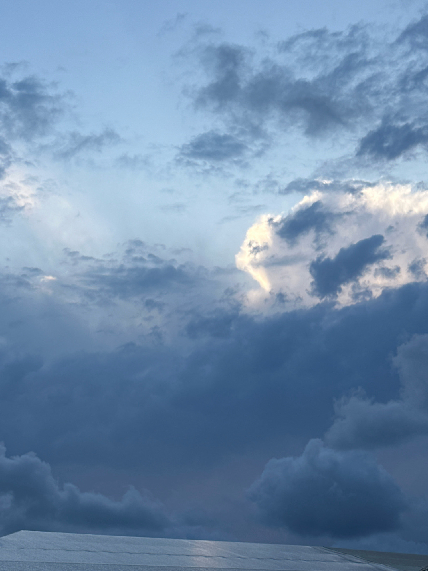 The image depicts a cloudy sky over a flat, reflective surface, possibly a rooftop or a body of water, in Lermontovo, Russia. The sky is a mix of light blue and dark gray clouds. Some clouds are illuminated by sunlight, creating bright white patches, while others are darker and more ominous. The lower part of the image shows a textured, light-colored surface that reflects the sky. There are no people or visible structures other than the implied surface beneath the sky. The overall impression is a natural landscape scene with dramatic cloud formations.
