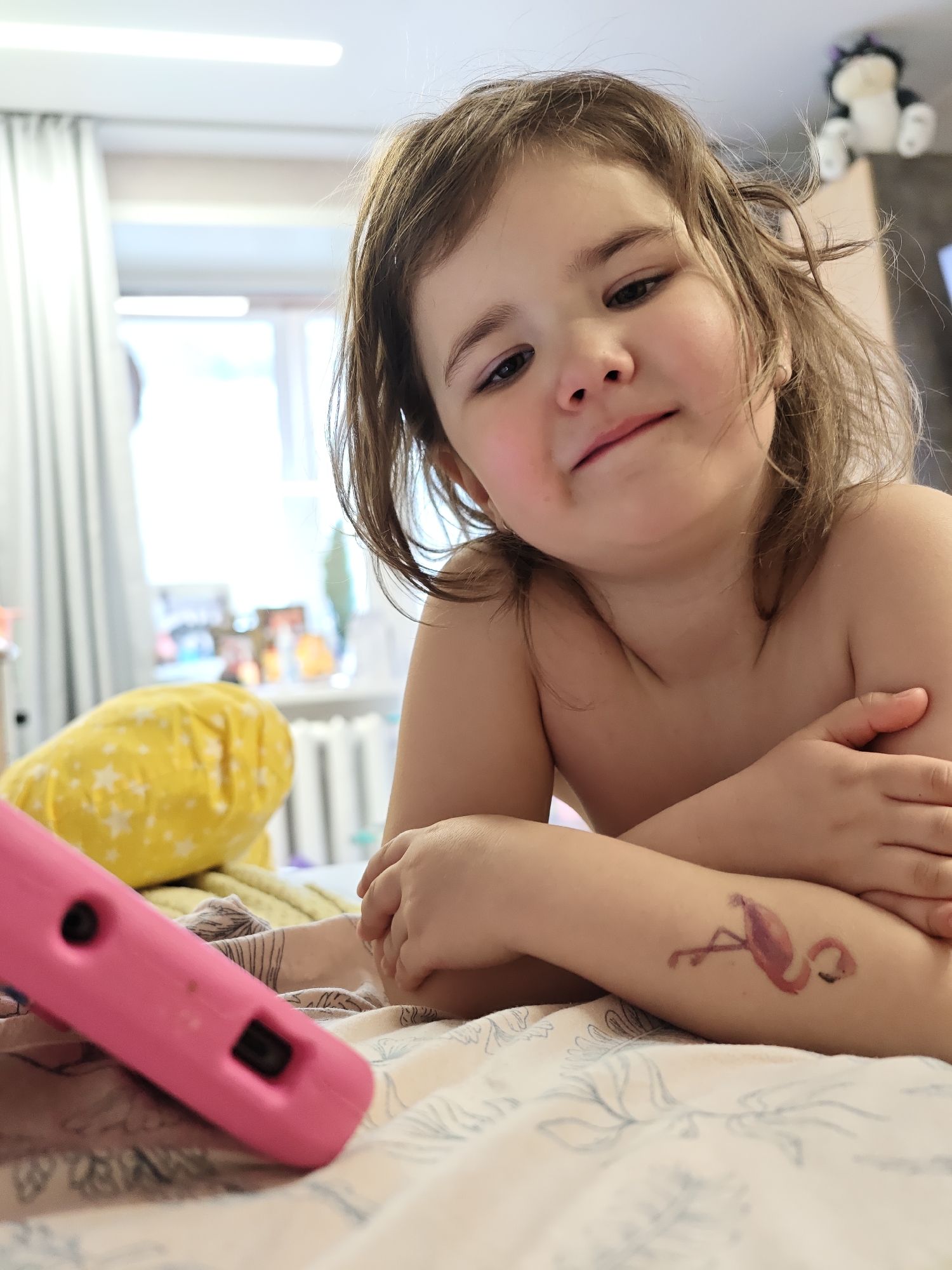 A young girl is lying in a bed, possibly in a private residence, given the casual attire. She is resting her arms crossed in front of her, her shoulders bare. She appears to have a temporary tattoo of a flamingo on her arm. There is a pink tablet on the bed beside her. The background shows a room with a window and light curtains. There is also a yellow pillow with white stars and a radiator behind the girl. The location is Chaikovsky, Russia, inferred from the request context.