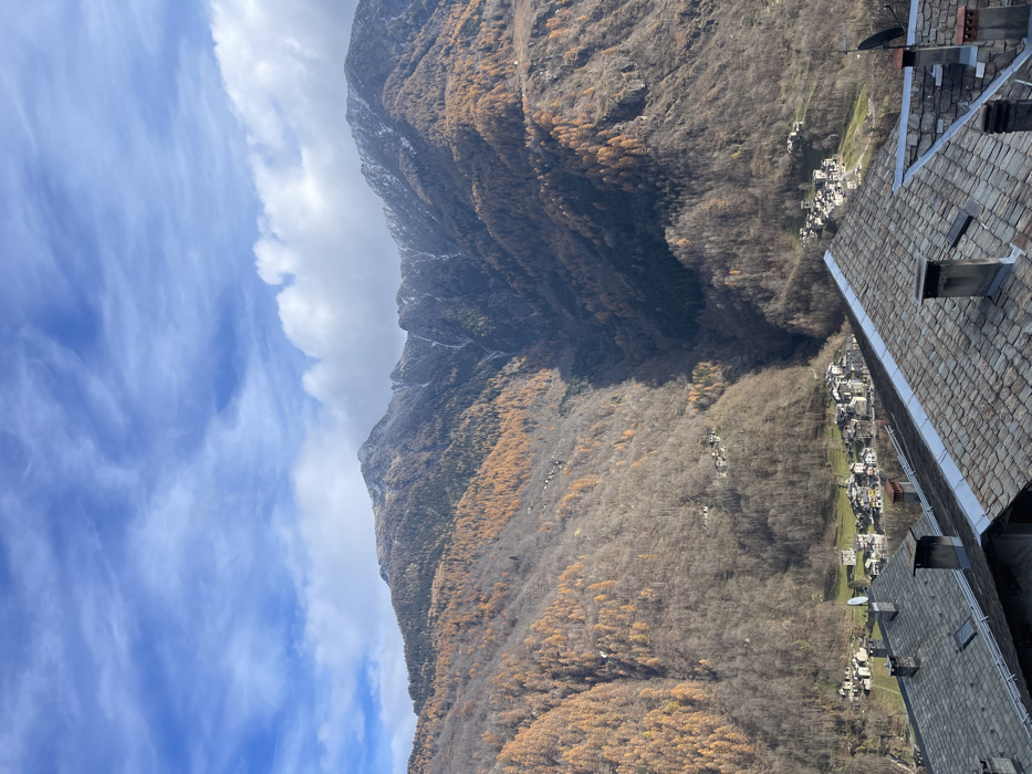 This image presents a stunning, wide-angle view of a mountainous landscape, likely captured from a high vantage point overlooking a valley in Dosso (Ciappanico), Italy. The vibrant blue sky, adorned with scattered, wispy white clouds, fills the upper portion of the frame.

Dominating the scene is a vast mountain range, its dark, rugged slopes covered in dense tree growth. Many trees appear to be deciduous, having lost their leaves, resulting in a muted brown and grey appearance across much of the terrain. However, striking patches of golden-orange foliage, likely larch trees, are visible along the mountainsides, indicating late autumn or early winter. Some higher peaks show hints of snow or frost.

A deep valley carves through the landscape, and nestled within it, a small village or collection of hamlets can be seen. The buildings are tightly clustered and feature traditional grey roofs. In the immediate foreground, on the right side of the image, the sloped grey slate roofs and chimneys of several buildings are prominent, suggesting the photograph was taken from an elevated position within or adjacent to this village. A satellite dish is visible on one of these foreground roofs.

The scene is bathed in bright daylight, with clear skies and visible shadows, enhancing the textures of the natural environment and the man-made structures. There are no people visible, and no specific activities are taking place, creating a tranquil, expansive landscape portrait. No visible text is present in the image.