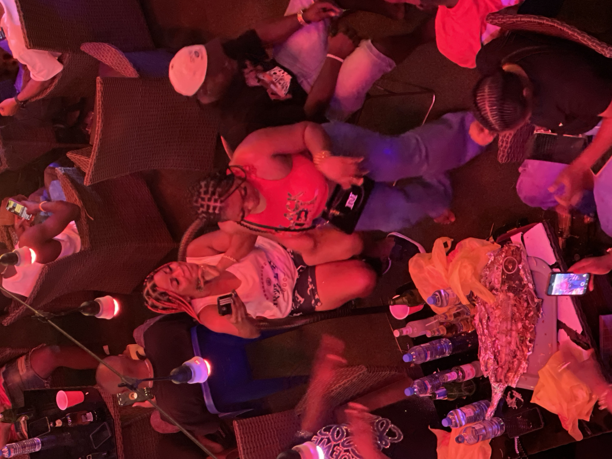 In an outdoor setting with red lighting, a group of people are gathered around tables laden with food and drinks. The scene appears to be a social gathering, possibly a restaurant or bar, given the presence of multiple tables, chairs, and various beverages. A large, foil-wrapped fish is a central element on one of the tables, suggesting a meal is being shared. Several individuals are holding phones, some of which are illuminated, indicating they might be taking pictures or videos. The overall atmosphere is casual and celebratory, with people dressed in relaxed attire.