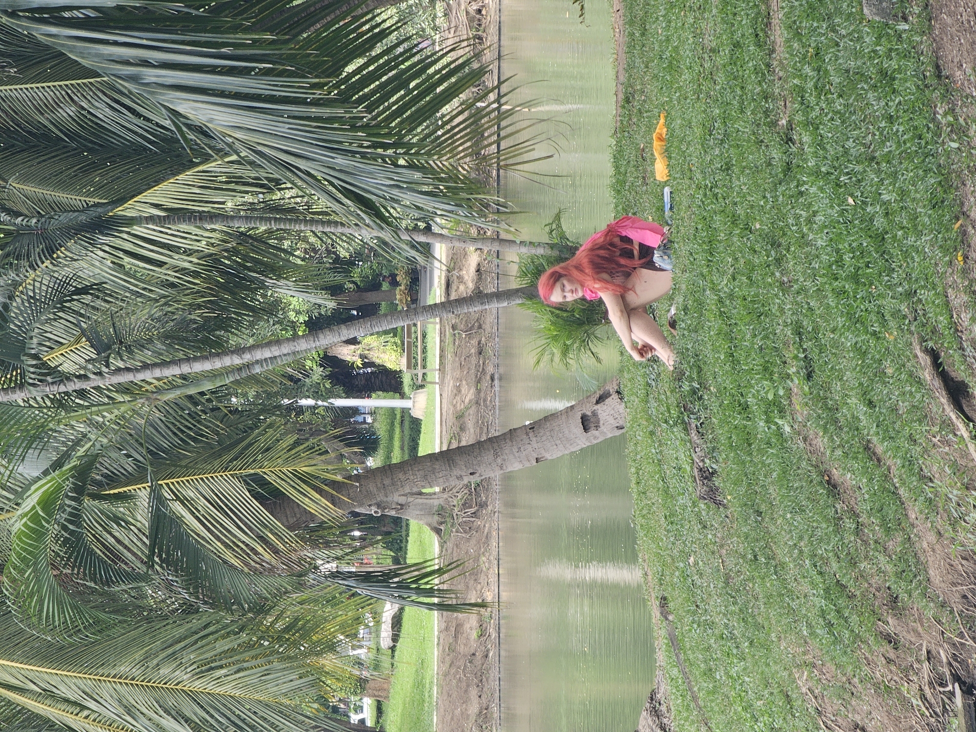 A young woman with long, bright red hair is sitting on a grassy embankment next to a body of water. She is wearing a pink top with a black strap and dark shorts. Her legs are crossed, and she is resting her arms on her knees, looking slightly down. To her right, on the grass, are a yellow cloth, a blue object that appears to be a phone, and a small dark pouch.
The setting is a park in Bangkok, Thailand, characterized by lush green grass and numerous palm trees with their fronds casting shadows. The water is calm and appears to be a pond or a slow-moving river, reflecting the greenery of the surrounding trees. In the background, across the water, there are more trees and some structures, including benches, suggesting a public park area. The overall atmosphere is tranquil and natural.
