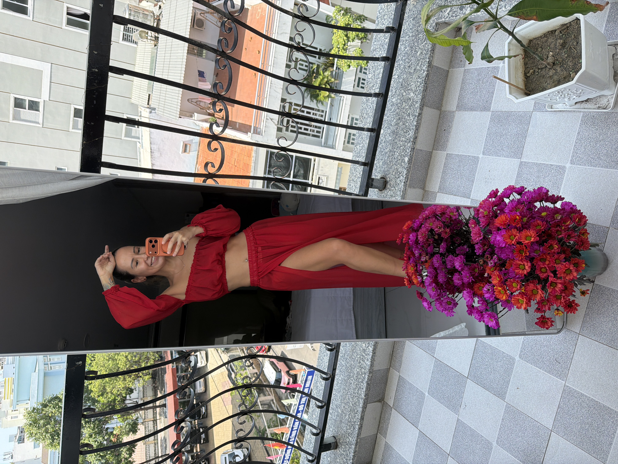 A woman in a red off-the-shoulder crop top and a long red skirt with a thigh-high slit stands on a balcony, taking a mirror selfie. She is smiling and holding an orange smartphone. A bouquet of colorful chrysanthemums in shades of pink, purple, and orange is in a vase on the floor in front of the mirror. The balcony overlooks a street in the city of Nha Trang Ward, Vietnam, with buildings and parked cars visible. The floor of the balcony is tiled in a gray and white checkered pattern. A potted mango plant is also on the balcony.