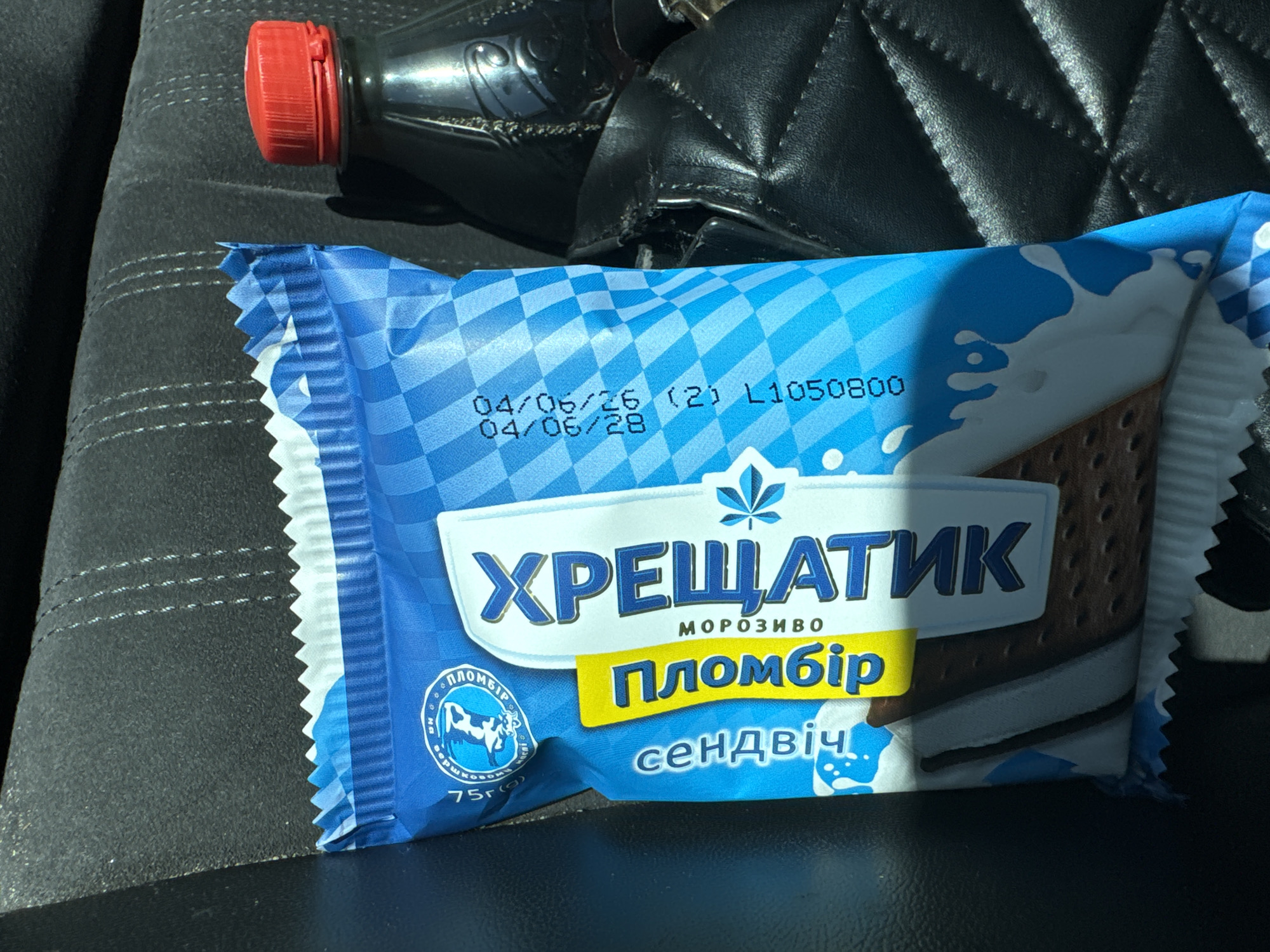 The image shows an ice cream sandwich package with Cyrillic text. The package is blue and white with a checkered pattern and graphics of milk splashes. The text on the package includes "ХРЕЩАТИК", "ПЛОМБІР", and "СЕНДВІЧ". There are also dates and codes printed on the package: "04/06/26 (21) L1050800". A red-capped bottle is visible to the right of the ice cream package. The background appears to be the interior of a vehicle, possibly a car seat made of dark textured material. The lighting suggests an outdoor environment, with sunlight casting shadows. The ice cream sandwich is the primary subject, positioned vertically in the frame. The location is identified as Sarny, Ukraine.