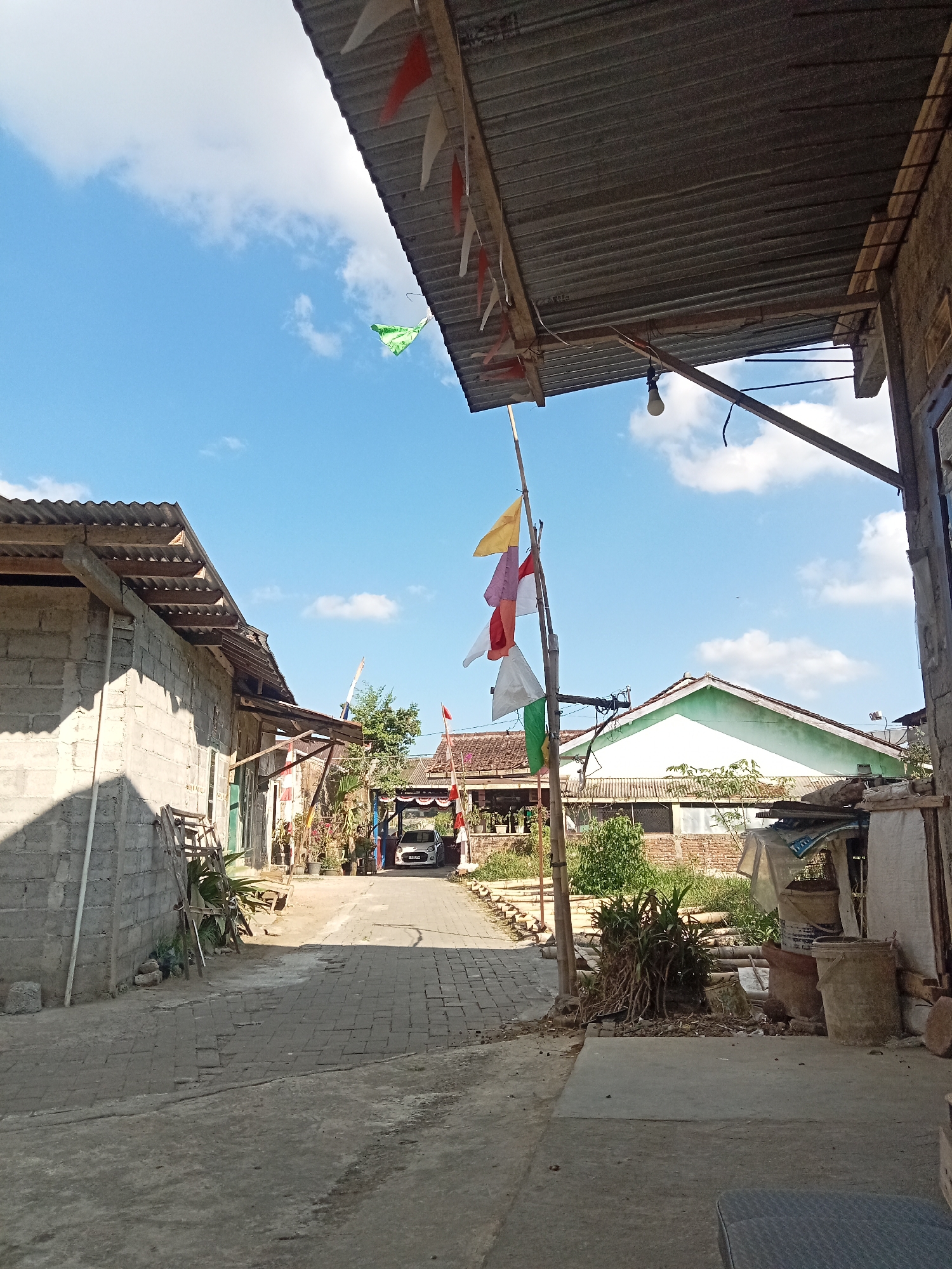 The image depicts a narrow, paved alleyway in what appears to be a residential area. The alley is lined with simple, low-rise buildings constructed from concrete blocks and corrugated metal roofing. In the foreground, to the right, a portion of an outdoor seating area with a dark textured surface is visible. A bamboo pole with several triangular flags, including red and white, and a green flag, stands prominently in the middle of the alley. The flags suggest a festive or celebratory occasion, possibly related to Indonesian Independence Day given the red and white colors.
The alley leads into the distance where a white car is parked. The buildings on either side of the alley are weathered and show signs of everyday use. On the left, a light-colored concrete block building casts a shadow across the ground. On the right, there is an assortment of objects, including buckets, plants, and what appears to be stored materials under a makeshift roof. The sky is blue with scattered white clouds. The overall impression is of a humble, working-class neighborhood in Pancurwening, Indonesia, decorated for an event. There are no people clearly visible in the image.