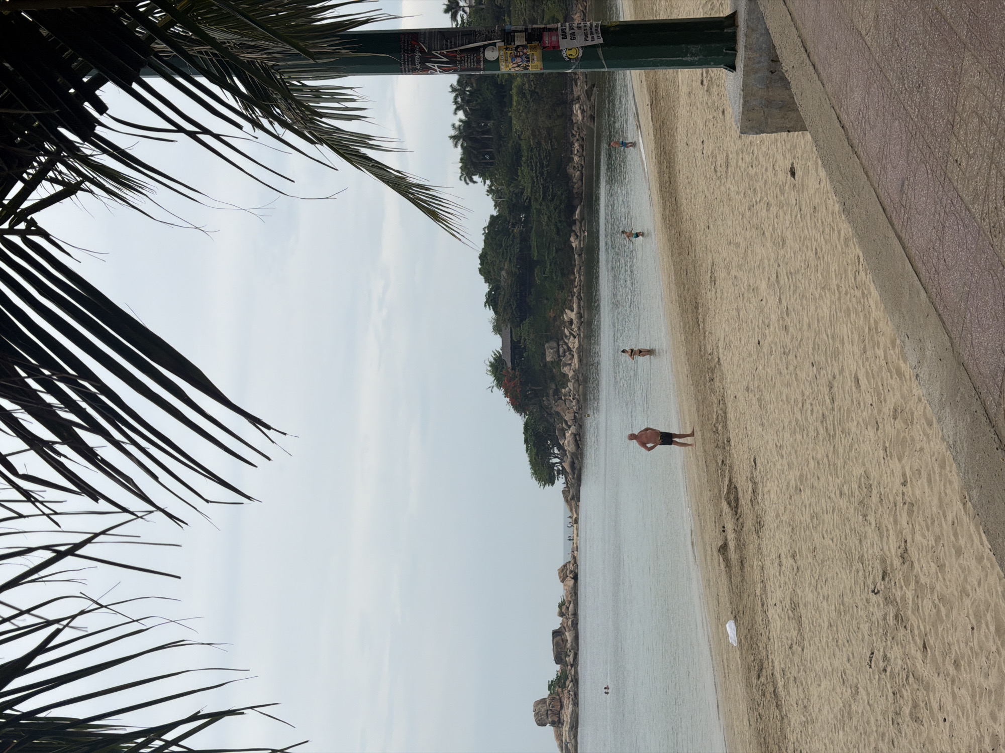 A beach scene photographed in Bac Nha Trang Ward, Vietnam, features a sandy shoreline beside a calm body of water. A man stands in the shallows near the shoreline, while a few other individuals are wading in the water further out. Lush green trees line the bank opposite the beach. Palm fronds overhang the left side of the frame, and a green pole with affixed flyers stands at the upper center. A paved path runs parallel to the beach along the right side.