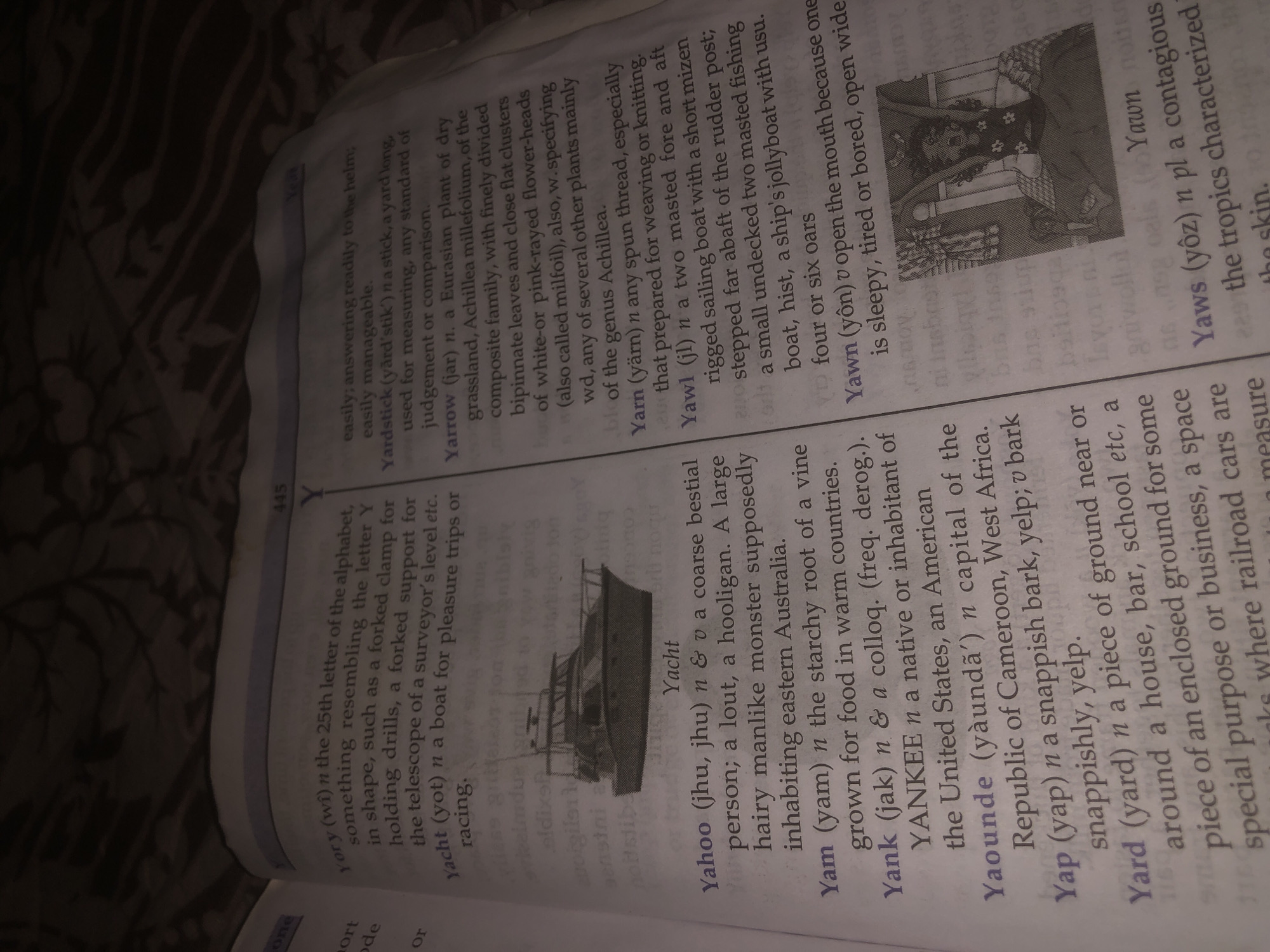 The image is a close-up of an open book, with text and an illustration. The book appears to be a dictionary, with entries for words such as "Yacht," "Yahoo," "Yam," "Yank," "Yaounde," "Yap," "Yard," and "Yawn." One entry, "Yacht," is accompanied by a black and white illustration of a yacht. Another entry, "Yawn," is accompanied by a drawing of a person yawning in bed. The text is in English, with some pronunciation guides. The page number "445" is visible at the top of the right-hand page. The background of the image is dark and textured, possibly a blanket or fabric. There is no discernible location information within the text or image that would connect it to Jalingo, Nigeria.