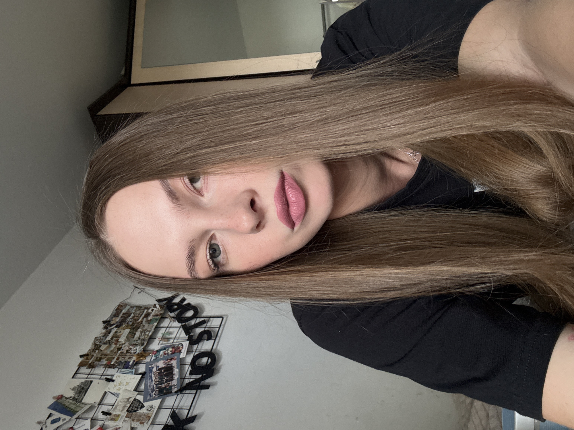 A young woman with long, straight, light brown hair is captured in a close-up, selfie-style shot. She is wearing a black t-shirt. Her makeup includes pink lipstick and subtle eye makeup. Her hair falls forward, obscuring part of her face. In the background, a black grid with various photos and postcards is visible on a white wall, along with black letter cutouts that appear to spell out "NO STORY." To the right of the woman, a portion of a wooden cabinet with a mirror is visible. The setting appears to be an indoor space, possibly a bedroom or personal living area. No specific location within Perm, Russia, can be determined from the visual information provided.