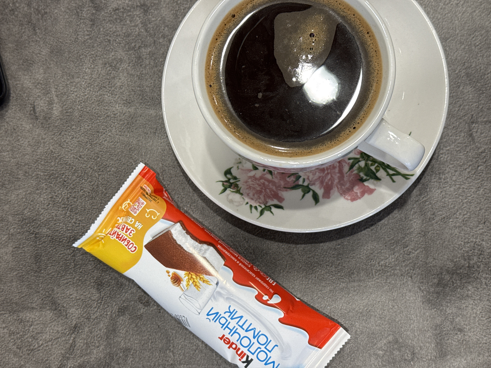 The image shows a cup of dark coffee on a saucer with a floral pattern, next to a Kinder Milk Slice snack. The coffee is reflecting light, and the Kinder Milk Slice packaging has Russian text. The objects rest on a gray textured surface.