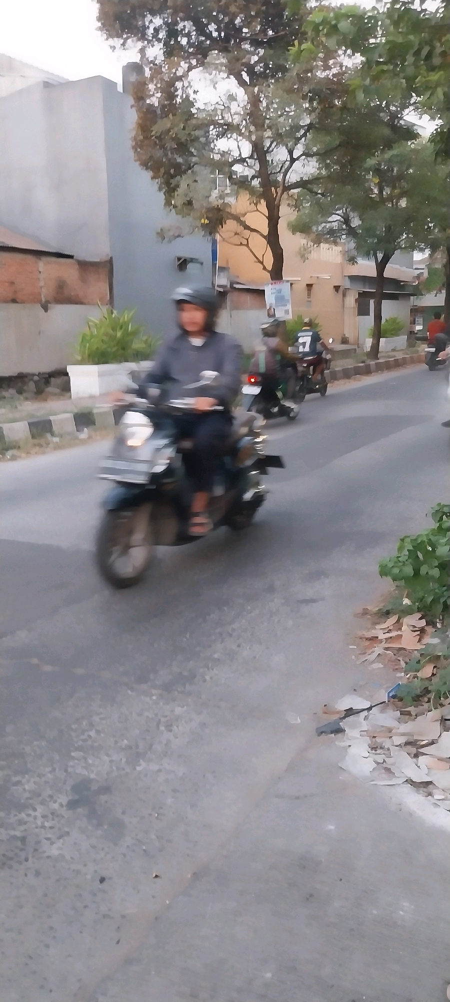 In Bojong Nangka, Indonesia, a person wearing a helmet and a grey jacket rides a dark-colored scooter. The scooter has a silver front fender and is in motion on a paved road. Behind this scooter, another person on a scooter is visible, wearing a dark top with a white number "7" on the back. Further down the road, a third scooter with a rider wearing red is partially visible. The environment includes trees with brown and green foliage, buildings with grey and brown facades, and a sidewalk with scattered debris. The lighting suggests it is daytime, possibly late afternoon or early morning.
