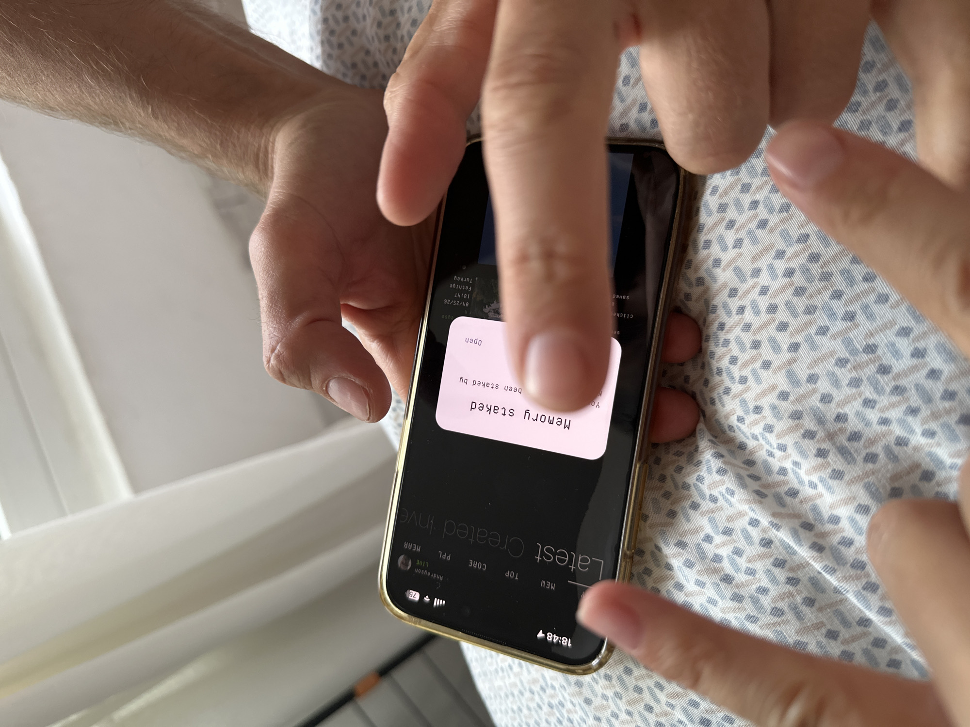 A person is holding a gold-colored smartphone displaying an app with the notification "Memory staked". The smartphone screen also shows the time as 18:48. The app appears to relate to investments, with "Latest Created Inve" visible. The screenshot displays 04/25/26 18:47 in Fethiye, Turkey. The individual's fingers are on the screen, suggesting interaction with the device. The person is wearing a patterned shirt.
