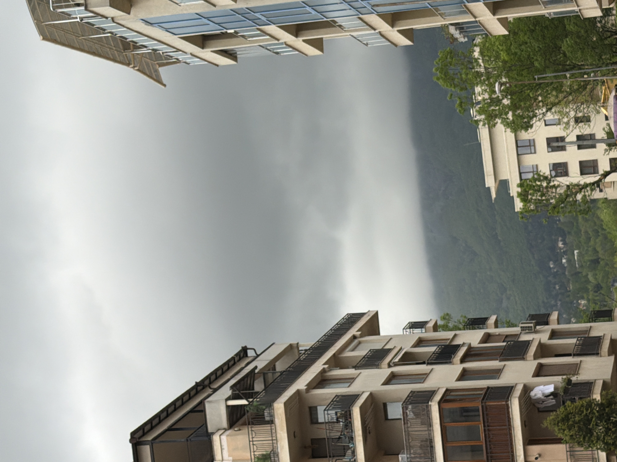 The image depicts a view of Sofia, Bulgaria, on an overcast day. Two apartment buildings frame a backdrop of mountains shrouded in mist and low clouds. The building on the top has a modern design with a geometric facade and glass windows. The building on the bottom is also residential, featuring balconies with iron railings and sun shades. The sky is uniformly gray, suggesting heavy cloud cover or impending rain. Vegetation is visible on the mountains and in some of the balconies, adding a touch of green to the otherwise muted color palette.