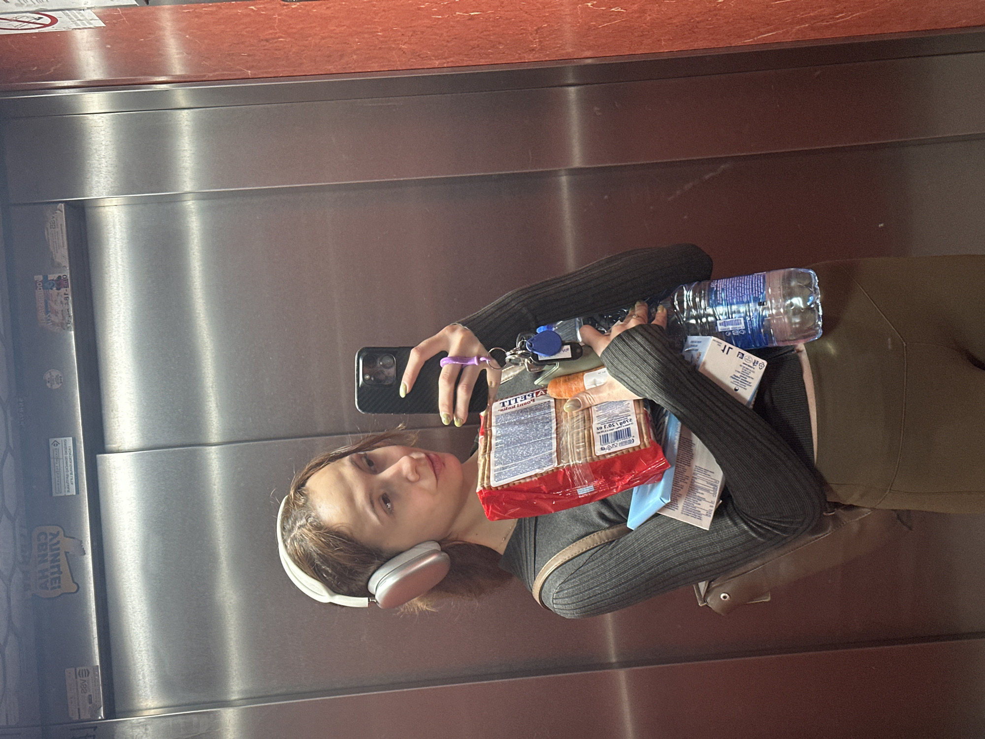 A young person, appearing to be female, is depicted in a selfie taken inside an elevator, located in Novi Sad, Serbia. She has brown hair, wears white over-ear headphones, a dark grey ribbed long-sleeve top, and olive-green bottoms. Her left hand holds a black smartphone, reflecting her face, and a set of keys with a blue tag and other trinkets attached to a purple strap. Her nails are painted a light green.
In her right arm, she balances a clear plastic bottle of water and several packaged items. The most prominent package is a red and white bag of crackers, clearly displaying the brand name "TITELLA" and the Cyrillic word "КРАКЕРЫ" (krakery, meaning crackers), along with "30 г" (30g). Other smaller, unidentified packages in blue and white are also visible. A brown shoulder bag strap is visible on her right side.
The setting is an elevator, characterized by its metallic, panelled walls, likely stainless steel, which reflect the interior. The top section of the elevator has a reddish-brown panel. On the left wall, several stickers or labels are visible, including a "NO SMOKING" sign at the top. Other visible text on the wall includes "DUNJA", "AUNTEO BUN HY", and "220V". The Cyrillic text on the cracker package is consistent with the location context of Novi Sad, Serbia. The person maintains a neutral to slightly relaxed expression. The lighting appears to be artificial indoor light.