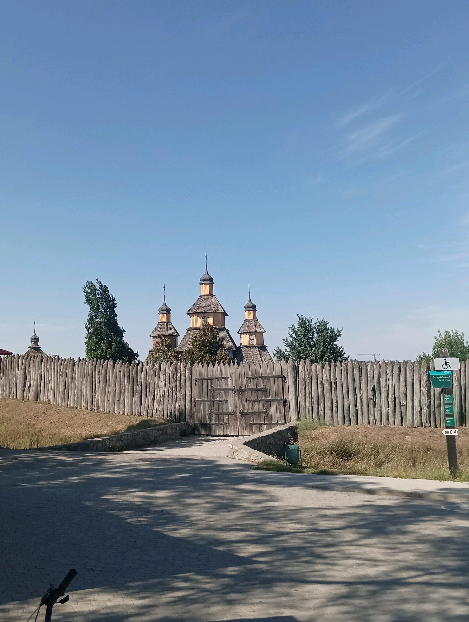 The image shows a historical wooden fort with a tall palisade fence and several wooden structures with onion domes. The fort is located in an outdoor environment with a clear blue sky and some wispy clouds. A paved path leads towards the fort's entrance, which is partially visible through a gate in the fence. There are trees and dry grass surrounding the fort. A signpost with directional arrows and an accessibility symbol is visible to the right of the fence. The overall scene suggests a preserved historical site or museum.