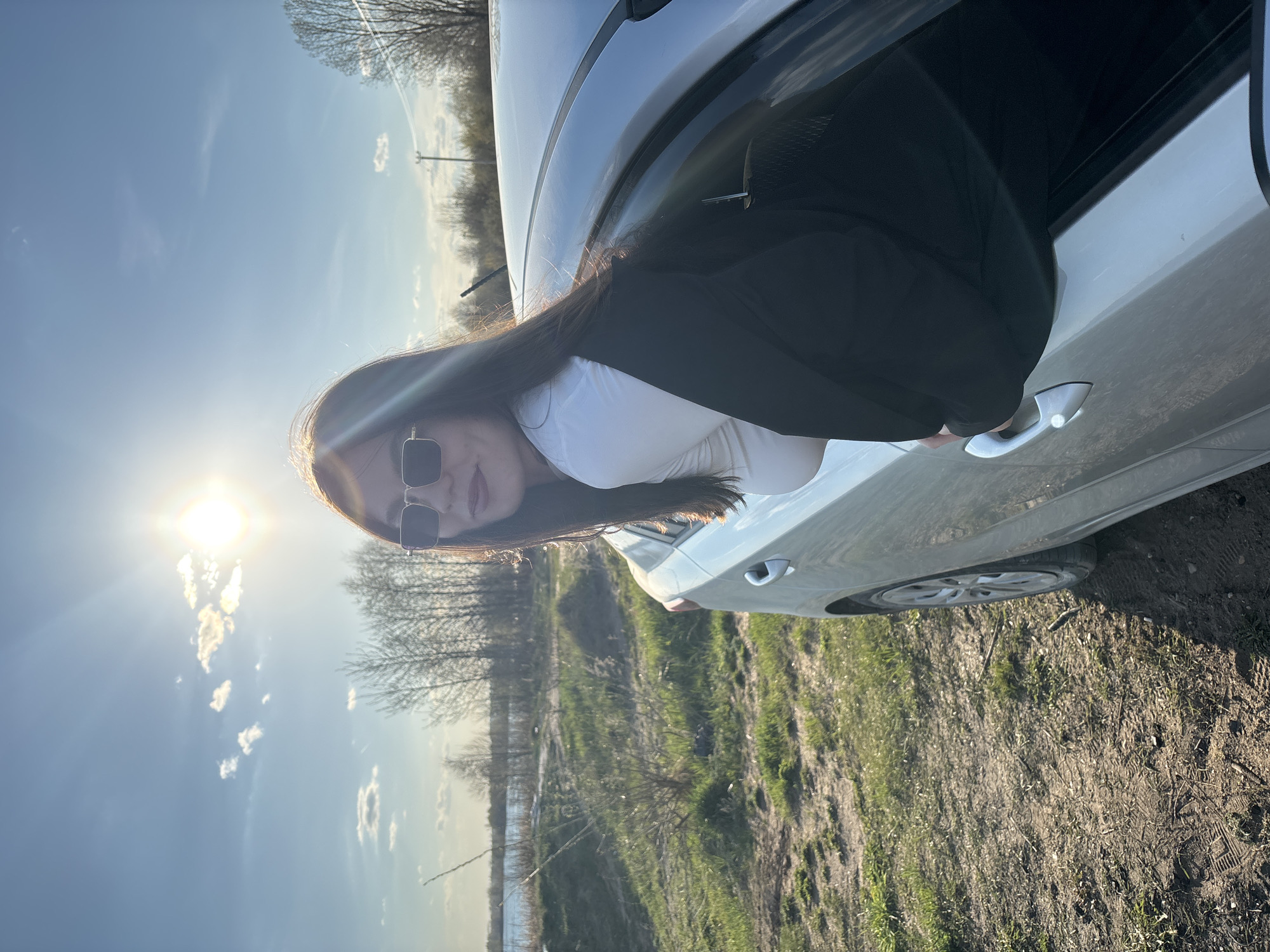 A woman with long, dark hair and sunglasses is sitting inside a light-colored car with her upper body leaning out the window. She is wearing a white top and a black blazer. The car is parked on a grassy field next to a body of water. The sun is shining brightly in the sky. The location is Zaosinovo, Russia.