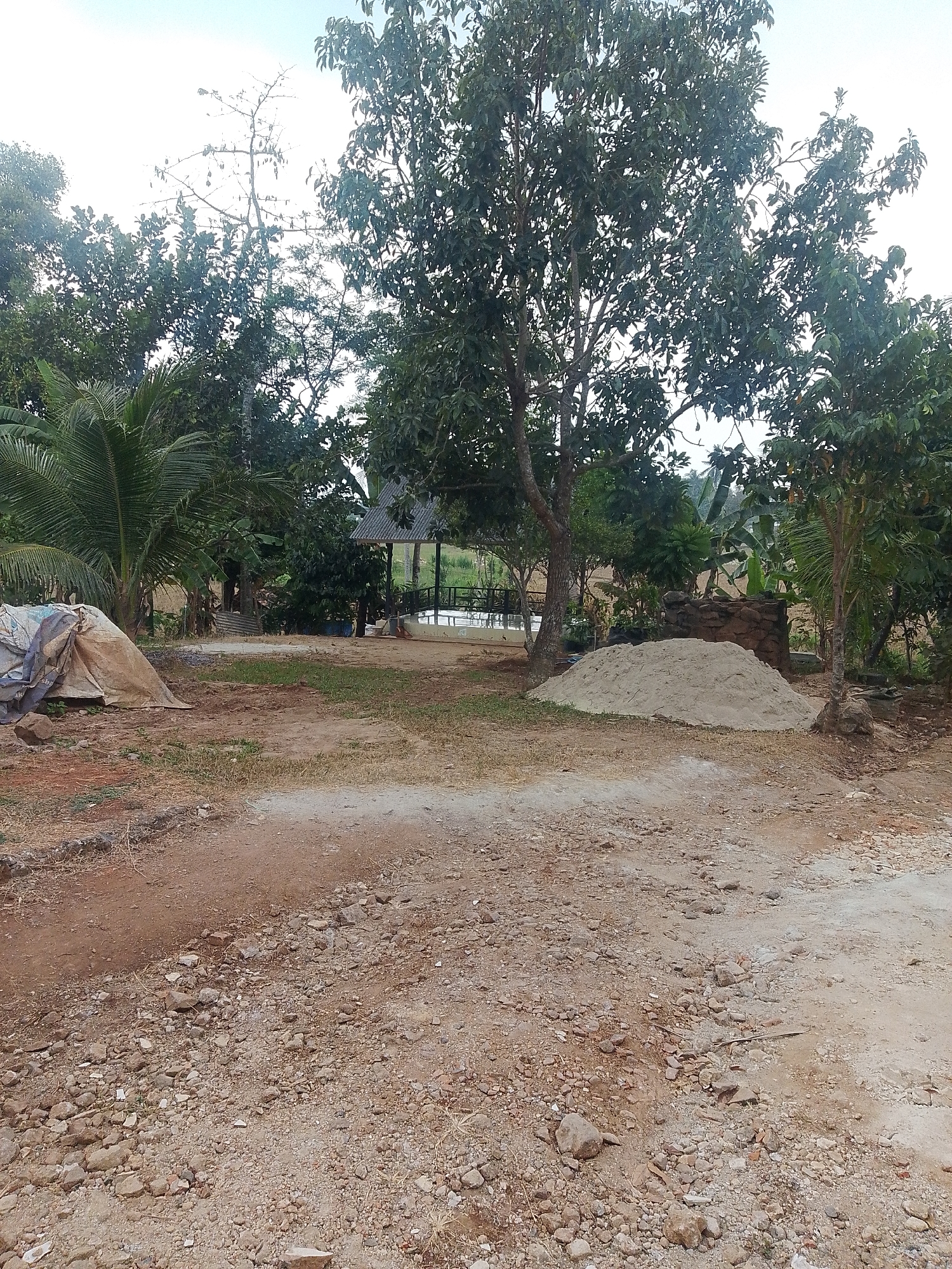 The image depicts an outdoor scene that appears to be a construction or landscaping site, possibly within the area of Kurungannyawa, Indonesia. In the foreground, a dirt path with scattered rocks and dry grass leads into the frame. To the left, a large tarp-covered object, potentially equipment or stored materials, rests on the ground. Further into the midground, a substantial pile of light-colored sand is visible. Behind the sand pile and to the right, a small, open-sided structure with a corrugated metal roof stands amidst lush green trees. The background is dominated by a dense canopy of various trees, including palms, with a bright, overcast sky overhead. There are no people visible in the image.