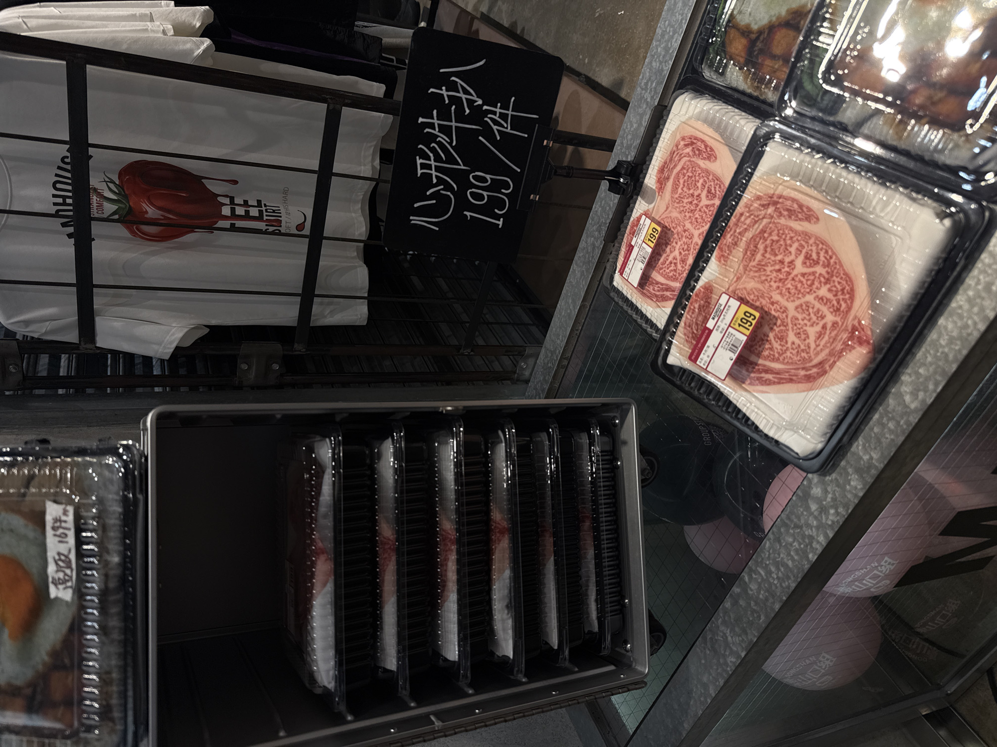 An overhead view captures an indoor retail setting, likely a supermarket or specialty store, featuring various products displayed for sale.
On the left, white t-shirts are arranged on a rack. One prominent t-shirt features a red, stylized, melting tomato-like design and text that includes "MADHOUSE," "COMFORT," "GROUP ST," "SUIRT" (likely "SHIRT"), "SOFT / 12HRS HARD," "DONGSHAN," and "LONGSHAN."
In the upper center, a black sign with white handwritten Chinese characters reads "心形牛扒" (Xīnxíng niúpá - Heart-shaped steak) and "199/件" (199 per piece/item).
To the right, several clear plastic containers hold cuts of highly marbled red meat, characteristic of premium beef. Two of these containers prominently display price tags marked "199" and the brand "MADHOUSE."
In the bottom left, another clear plastic container holds a food item, possibly a prepared meal or sushi, with a sticker visible that reads "高級 169円" (Gāojí 169 yuán - High-grade 169 Yuan/Yen). Adjacent to this, a metal display case or refrigerator section contains multiple vertical slots, each holding additional clear plastic containers, likely containing more meat or food products.
The reflective floor below the displays shows faint reflections, including circular pink graphics with Chinese text such as "山口组" (Yamaguchi-gumi), "DONGSHAN," and "LONGSHAN." The overall scene suggests a modern grocery or food market setting.