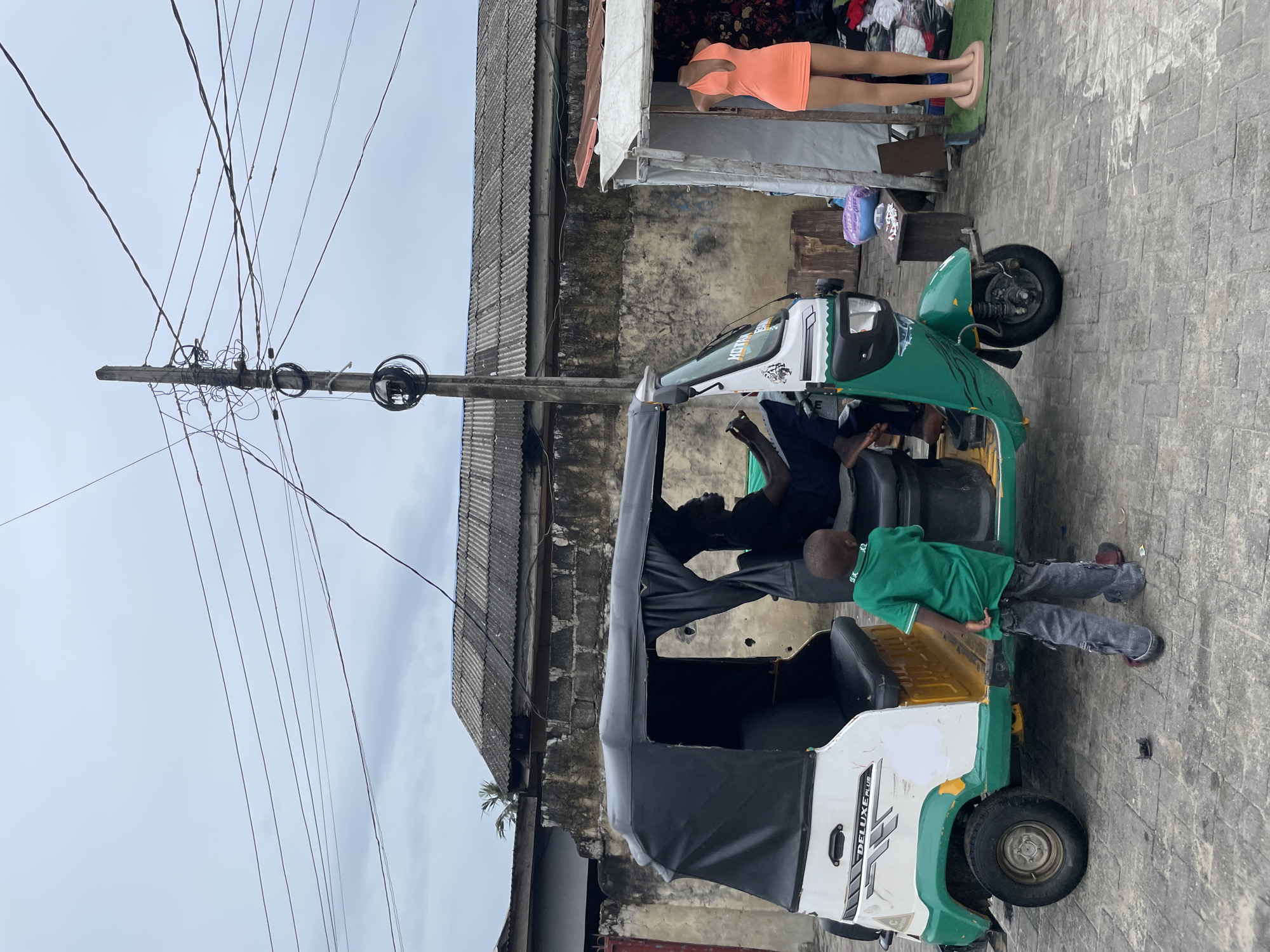 The scene depicts a three-wheeled vehicle, likely a "keke" or auto-rickshaw, parked on a paved street in Ado, Nigeria. A person is seated inside the vehicle, and another person is leaning against the side. Overhead, a wooden utility pole is densely strung with power lines. Adjacent to the vehicle is a building constructed of corrugated metal and cinderblock. A mannequin in an orange dress is visible in a window.