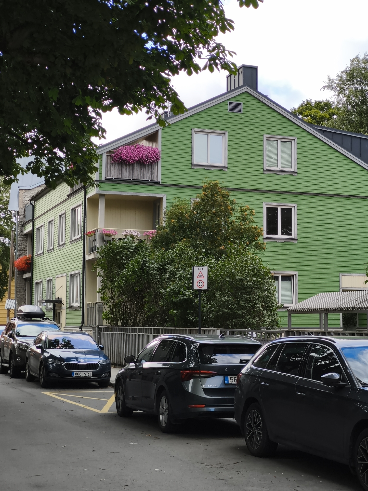 In Tallinn, Estonia, a bright green wooden building with multiple stories is captured in a street-level view. The building features white-framed windows and balconies adorned with abundant pink and red flowers. A dark-colored SUV with a roof box is parked in the foreground, followed by two dark-colored station wagons. The street is paved, and yellow parking lines are visible. A traffic sign indicating a speed limit of 30 is present. Lush green trees partially obscure the view of the building and the sky.