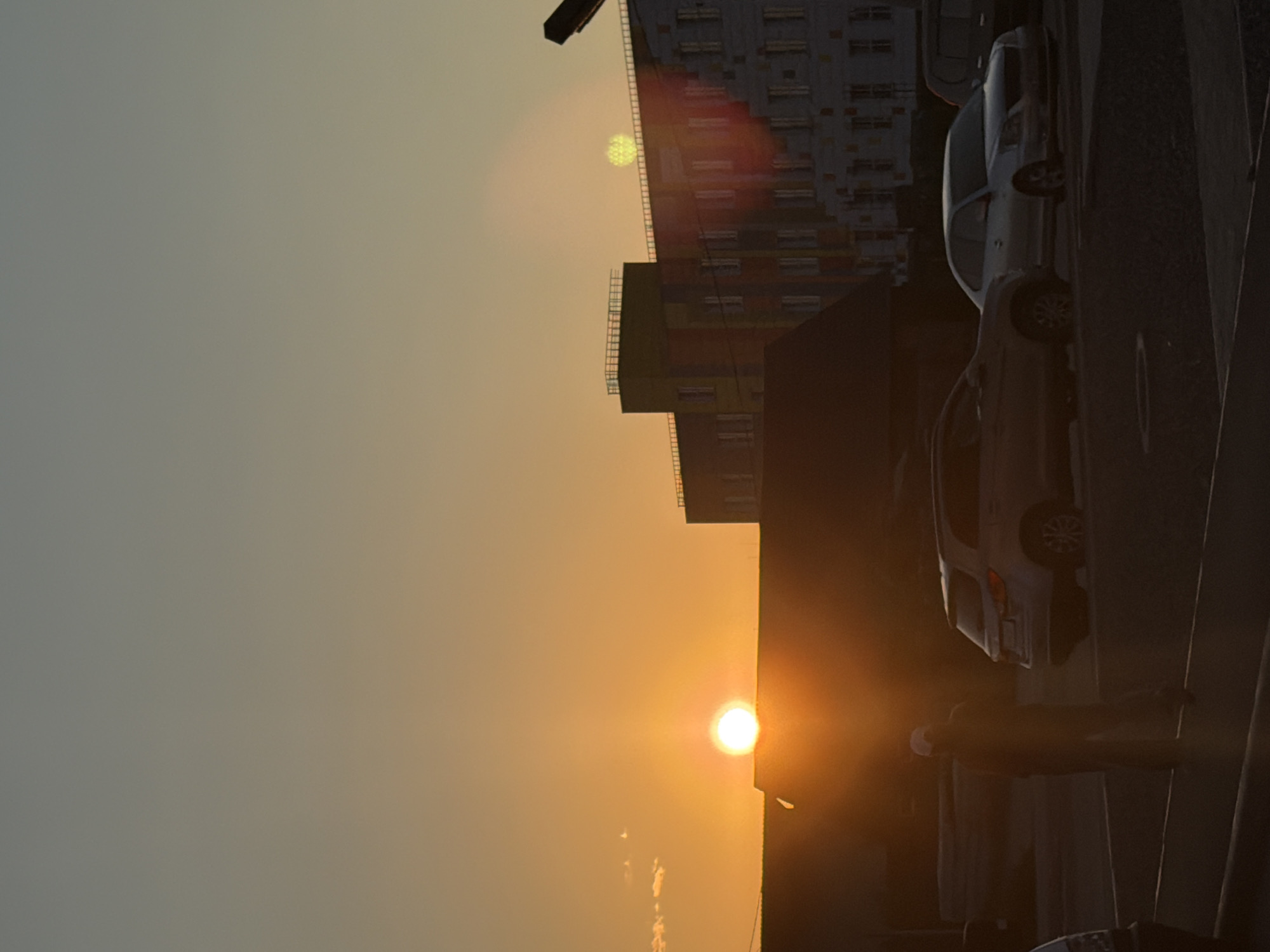 An outdoor shot captures a street scene in Kyzyl, Russia, during sunset. The sky is a gradient of orange and gray, with the sun positioned just above the horizon. In the foreground, a person wearing a white cap stands in the middle of a parking lot. Several cars are parked around the lot, including a silver sedan and a silver SUV. In the background, a large, multi-story building with colorful accents stands against the sky.