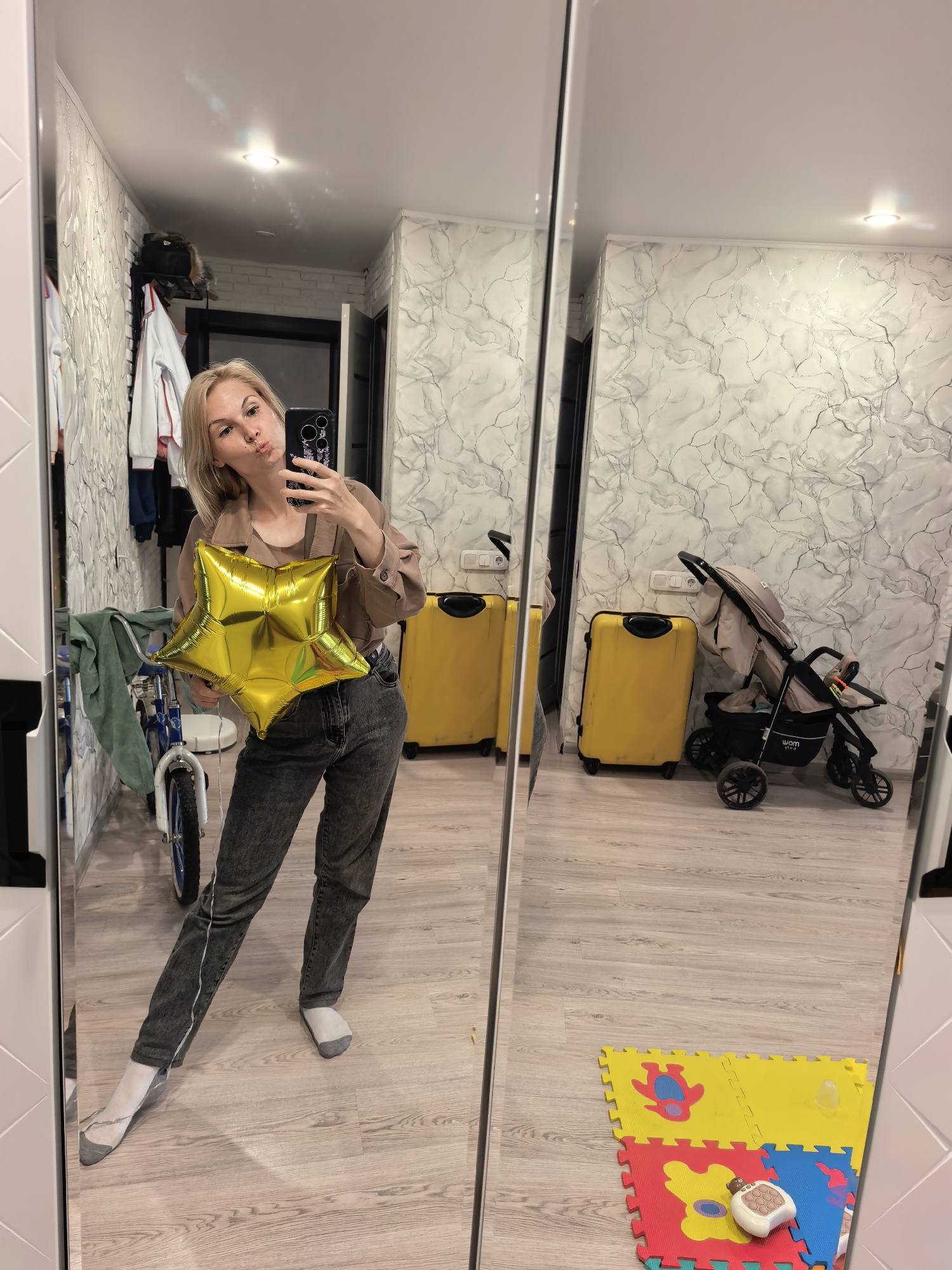 This full-length mirror selfie depicts a blonde-haired adult female, estimated to be in her 20s or 30s, making a pouting expression while holding a black smartphone in her right hand and a metallic golden star-shaped foil balloon in her left hand. She is dressed in a light brown jacket, a darker brown top, and dark grey jeans, wearing mismatched socks (light grey on the left foot, white on the right). The scene, located in Sterlitamak, Russia, reflects an interior space. To the left of her reflection, a clothing rack with various garments and a blue and white child's bicycle with green fabric draped over it are visible. To the right, two bright yellow hardshell suitcases and a light beige baby stroller with a black frame are positioned against a wall. The walls are covered in a light-toned, grey-patterned wallpaper resembling marble, with some silver accents. White electrical outlets are mounted on the wall. The ceiling is plain white with a single recessed light fixture. The floor is light wood or laminate. In the bottom right corner of the reflection, several interlocking foam playmats in yellow, red, and blue are laid out, with a white baby toy or remote on one of them.
